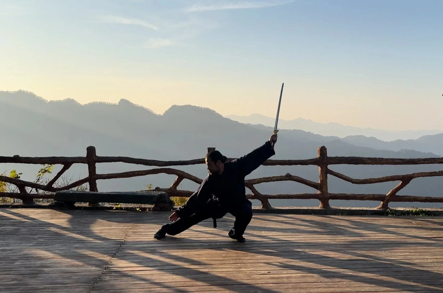 A person dressed in black practicing a martial arts pose with a sword on a wooden platform overlooking mountains during sunset.