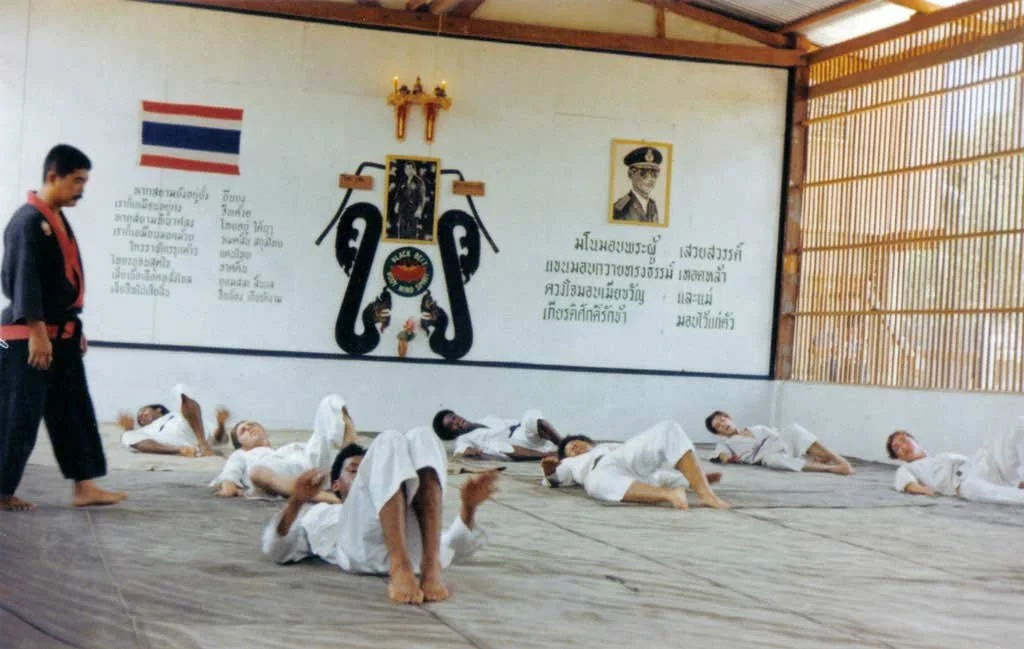 Students practicing martial arts on the floor of a dojo, with an instructor observing. The wall has a Thai flag, a portrait of a man in a military uniform, and a decorative symbol with black snakes, flags, and a flower.