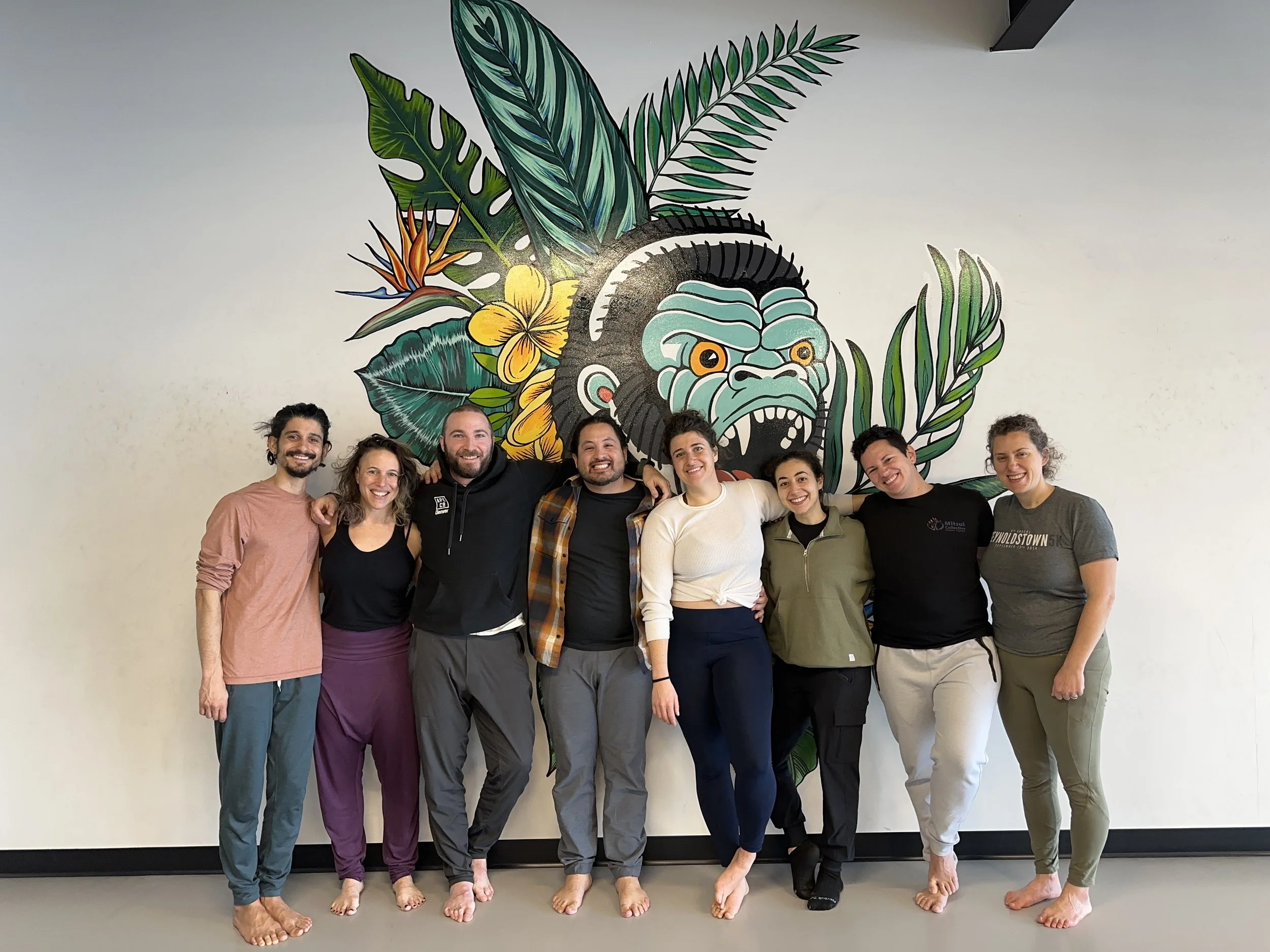 A group of eight diverse people standing closely together barefoot in front of a wall with a colorful jungle-themed mural of a gorilla face and tropical plants, smiling and posing for the photo.