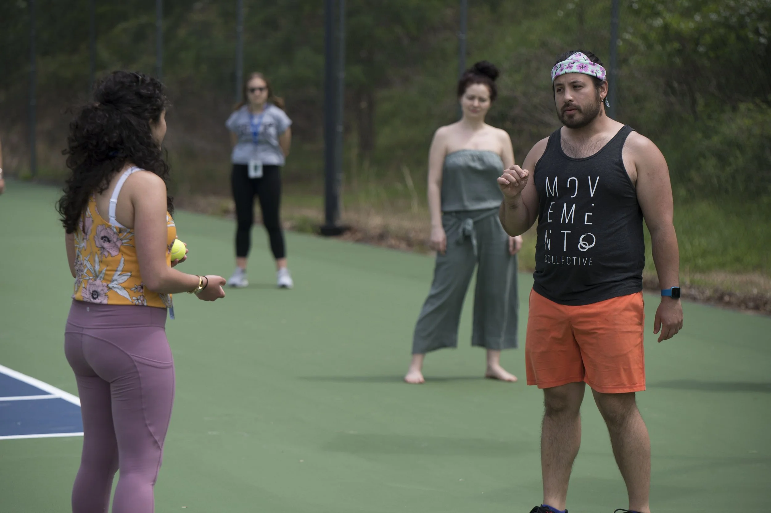 A group of people on an outdoor tennis court, with one woman holding a tennis ball and two women standing in the background, one in a towel dress and the others in casual clothing, engaging in conversation.