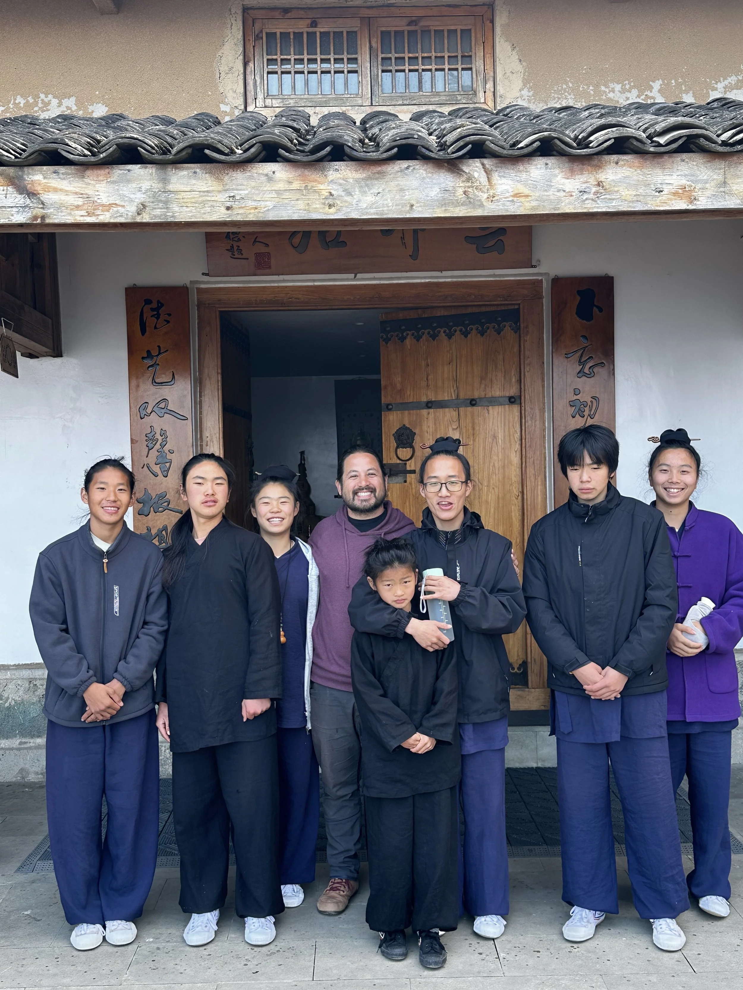 Group of seven people standing outside a building with traditional Chinese architecture, including a wooden door and tiled roof. The group is smiling and dressed in martial arts uniforms, with some wearing glasses and one child holding a water bottle.