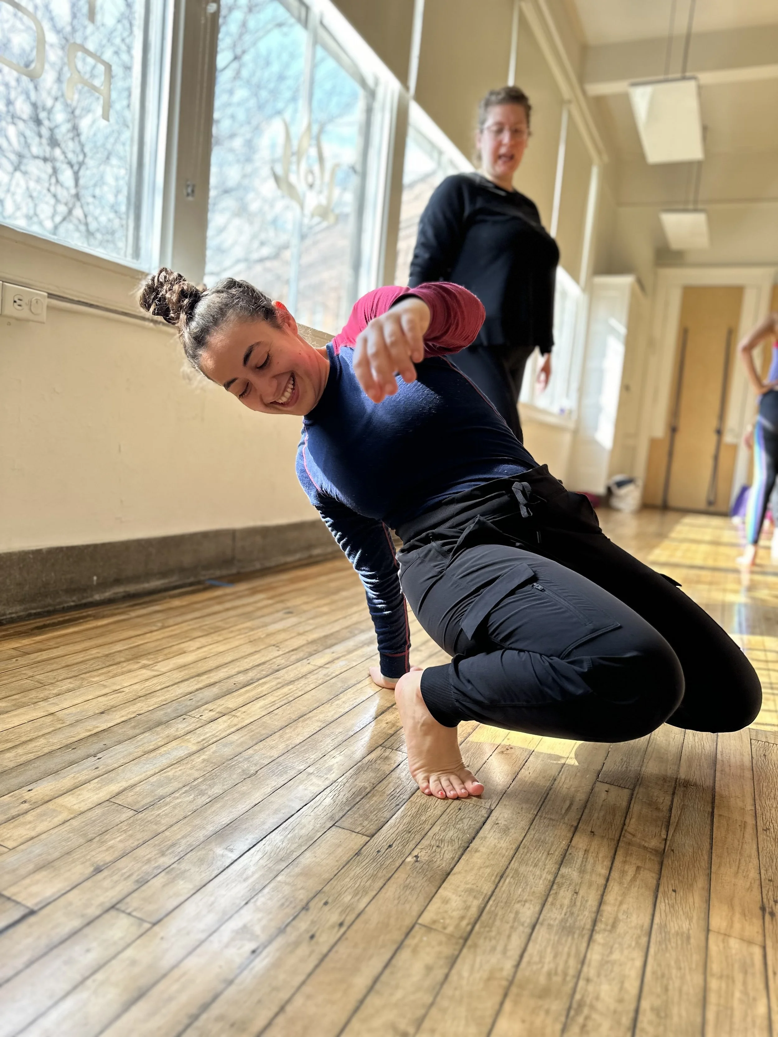 A woman practicing yoga indoors on a hardwood floor, balancing on one foot with the other leg crossed over her standing leg, and her hands on the floor. A woman in a black jacket stands behind her, observing or instructing. Large windows let in natural light.