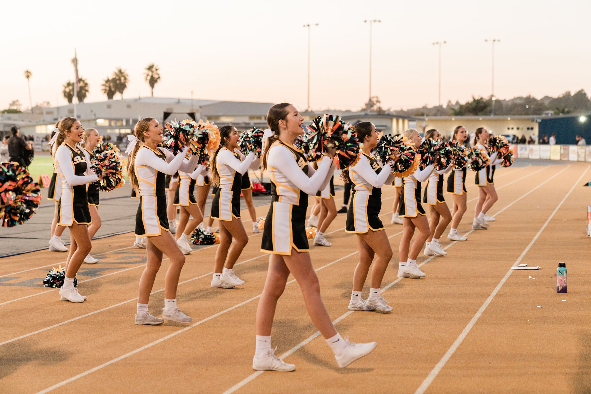 A group of cheerleaders in black, yellow, and white uniforms performing a dance routine on a track field at sunset.