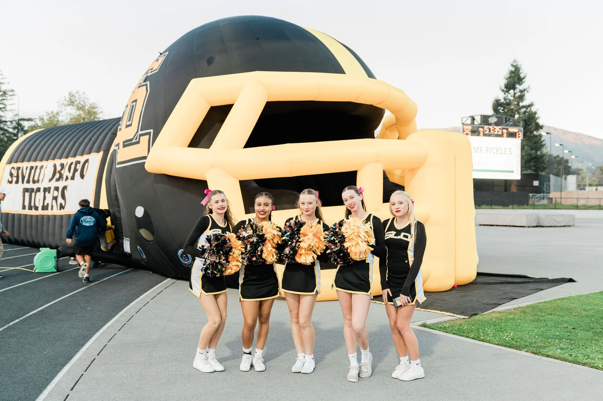 Five cheerleaders in black and gold uniforms smiling and holding pom-poms, standing in front of a giant inflatable football helmet with a black and yellow design at a sports stadium.