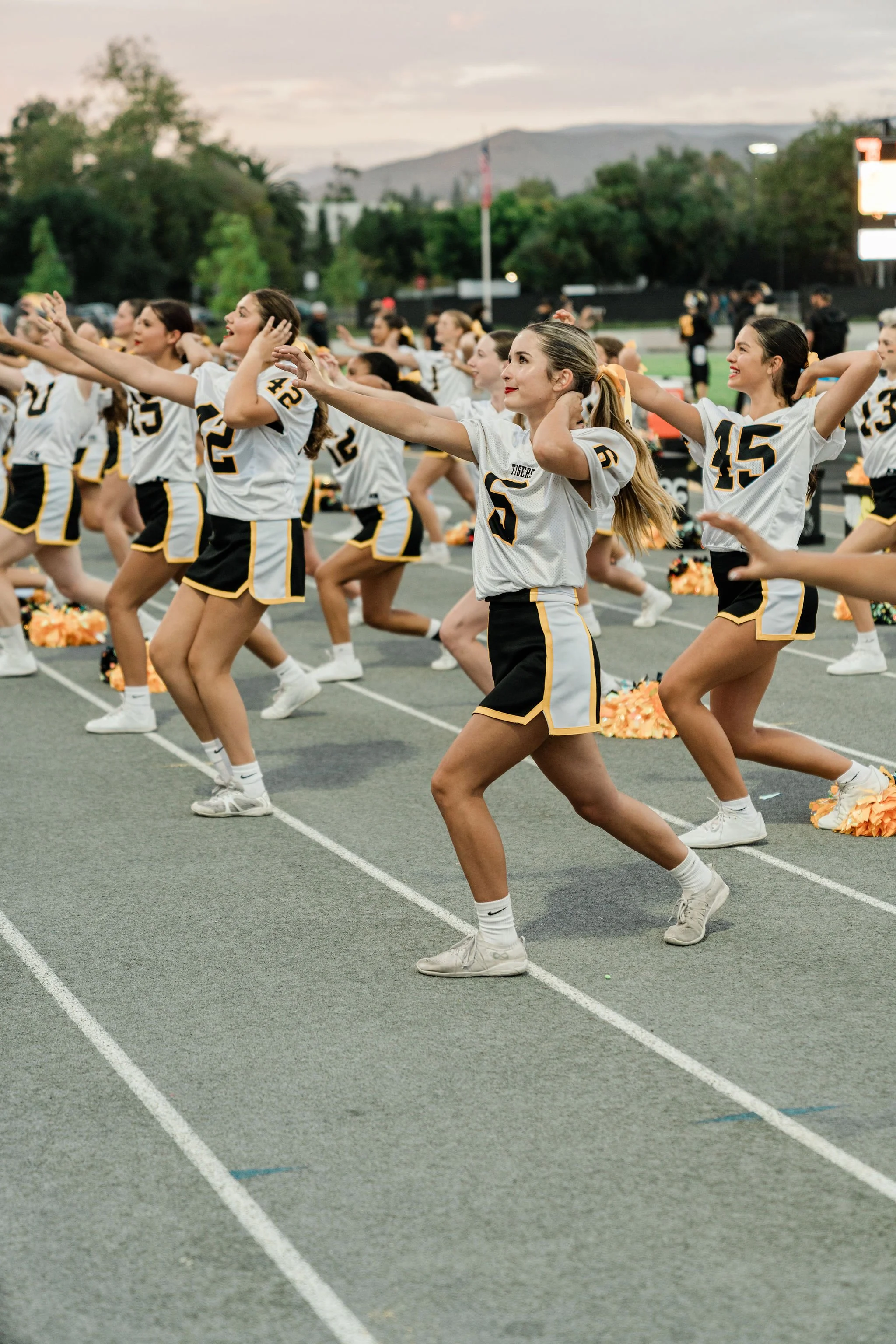 A group of cheerleaders performing a synchronized dance routine on a football field during the evening, wearing white and black uniforms with yellow accents.