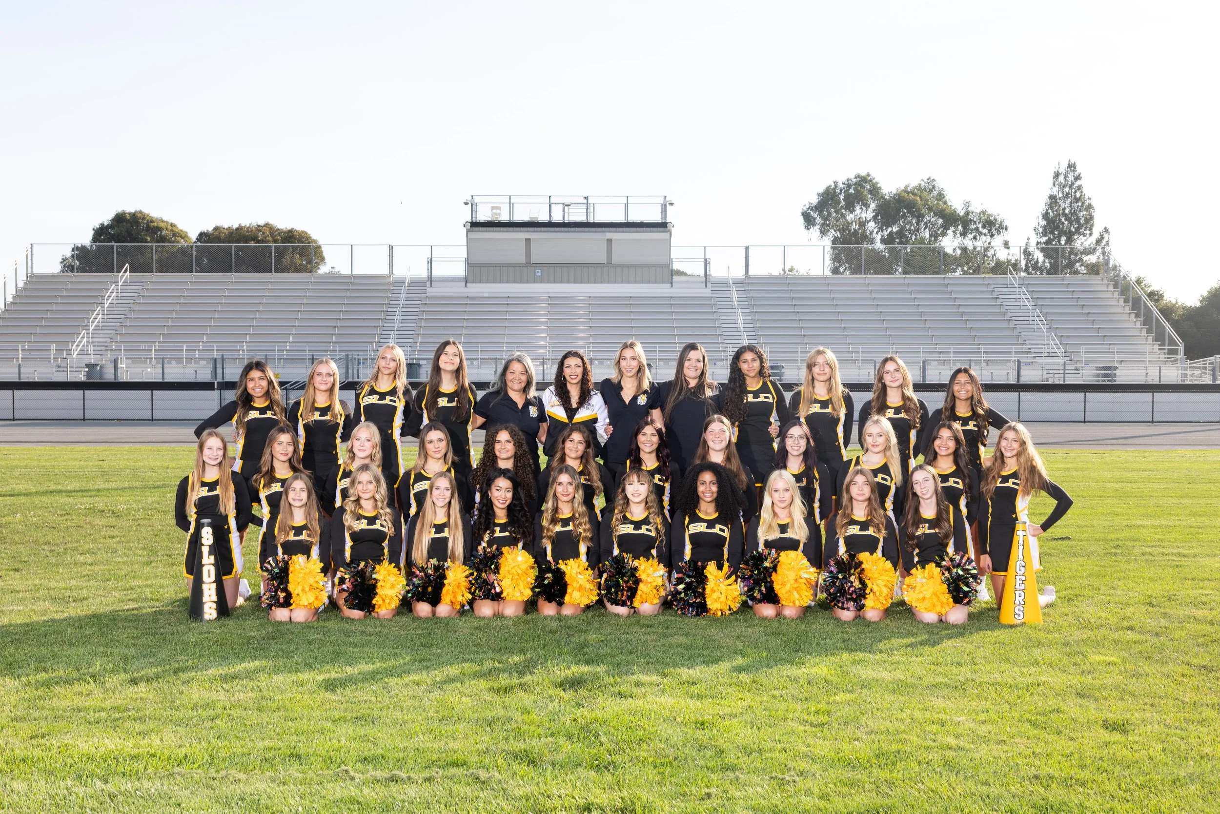 Group of cheerleaders in black and yellow uniforms standing on a football field with empty bleachers in the background.