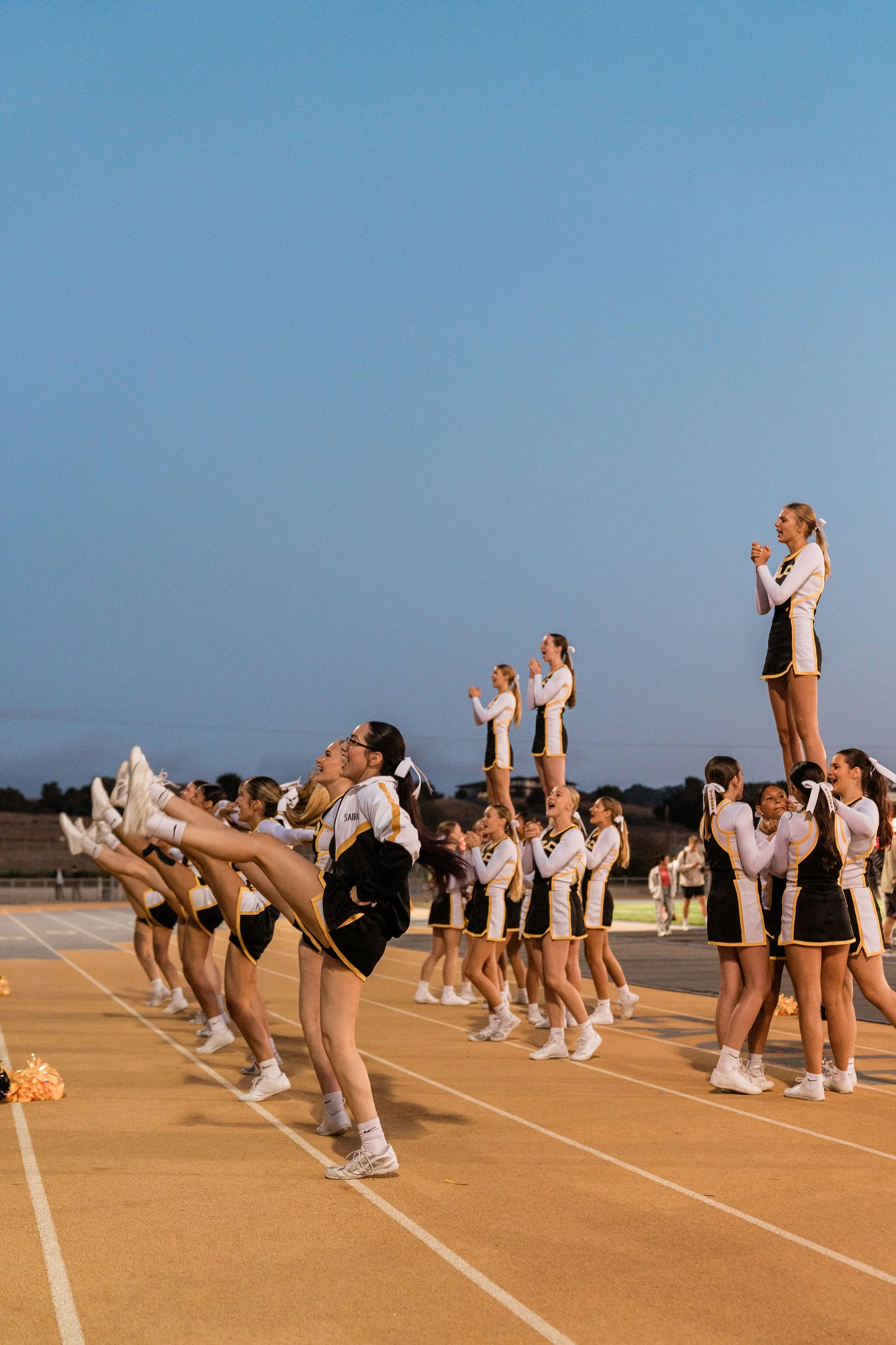 Cheerleaders performing a routine on a track field during evening, dressed in black and yellow uniforms, with a clear sky overhead.