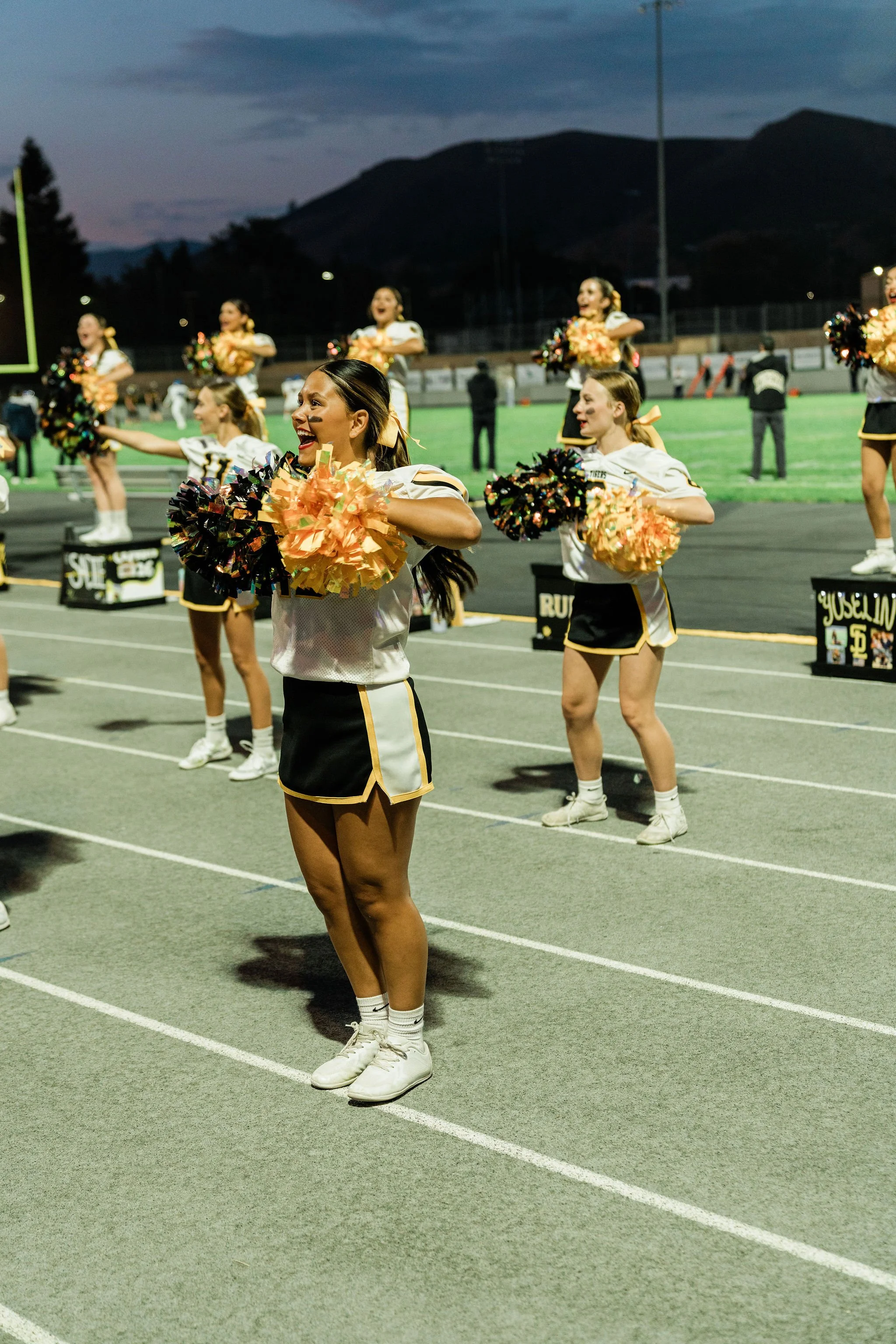 Cheerleaders performing on a football field at dusk, wearing white and black uniforms with gold accents, holding colorful pom-poms, with mountains in the background.