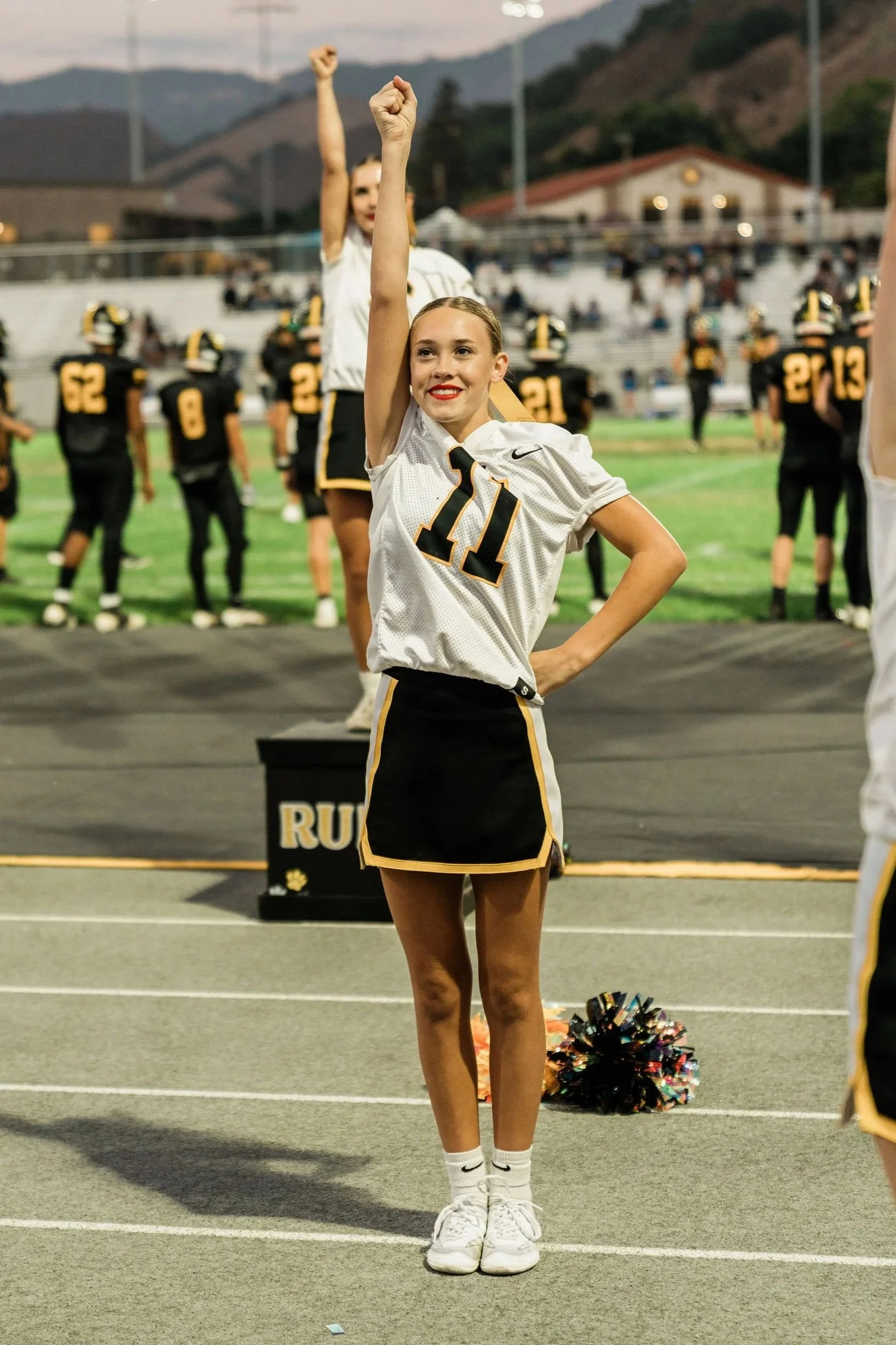 A cheerleader in a white uniform with the number 11 stands on a track with her right arm raised and a smile on her face. Other team members are behind her on a football field, and there are spectators in the bleachers.
