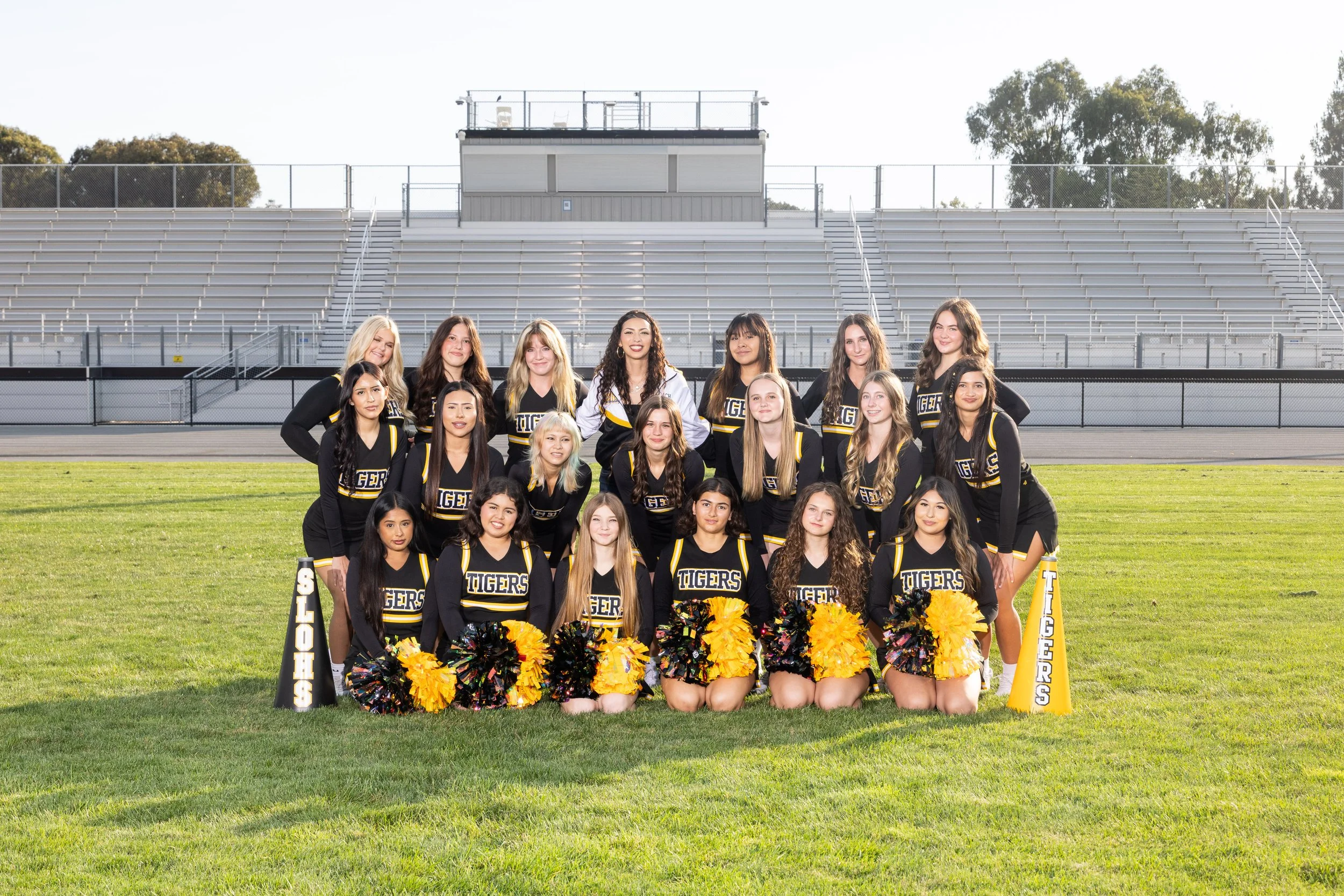 A group of cheerleaders in black and yellow uniforms with 'TIGERS' written on them, posing on a football field. Some cheerleaders are holding yellow and black pom-poms.