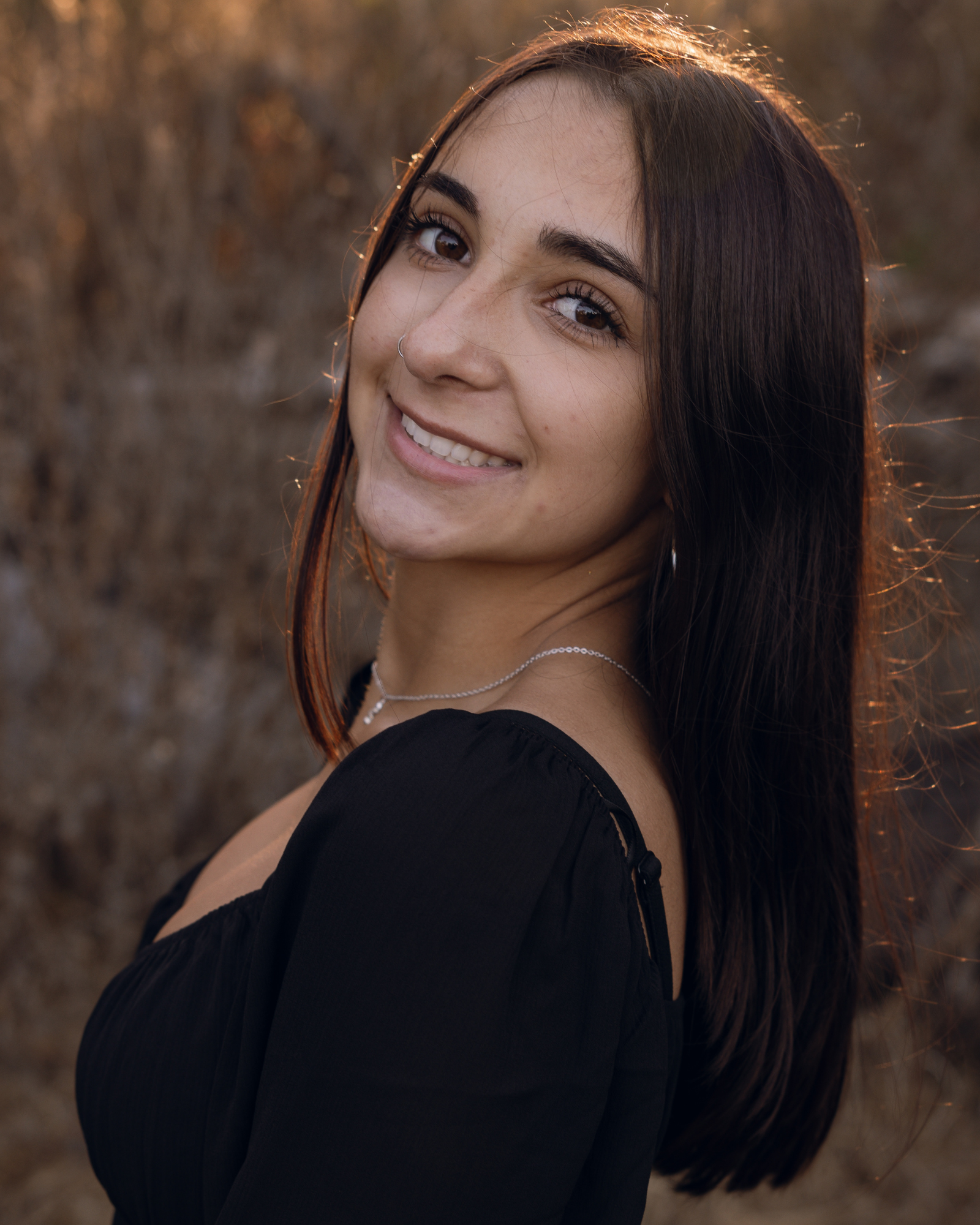 A young woman with long dark hair and a nose ring smiling outdoors at sunset, wearing a black top and a thin necklace.