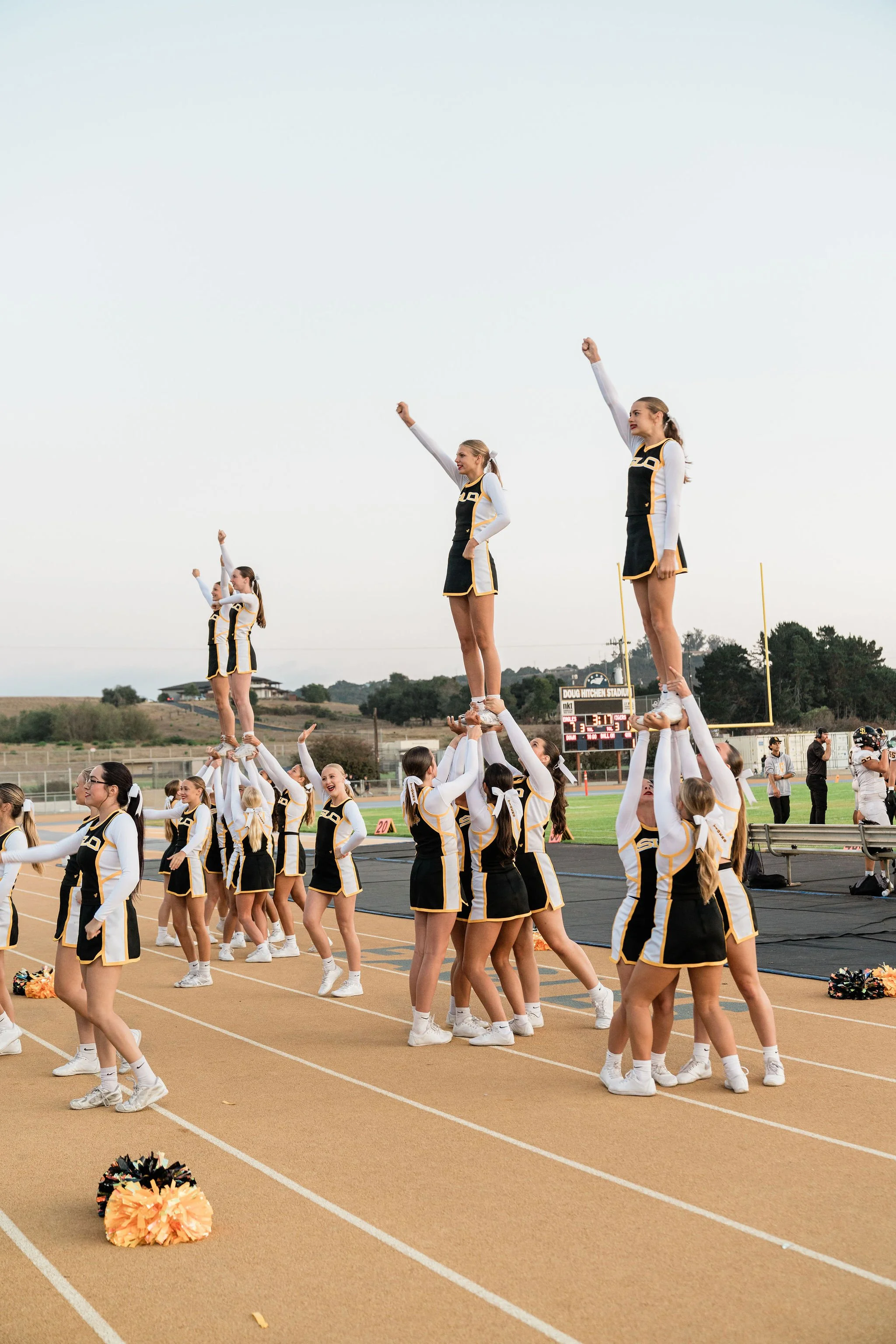 A group of cheerleaders performing a stunt on a track at a football field during daylight. Three cheerleaders are standing on the hands of their teammates, raising their fists in the air. They are dressed in uniform black and yellow outfits with white long-sleeve shirts underneath. Multiple other cheerleaders are smiling and walking in the background, with a football goal post and a scoreboard showing the game score and time in the distance.