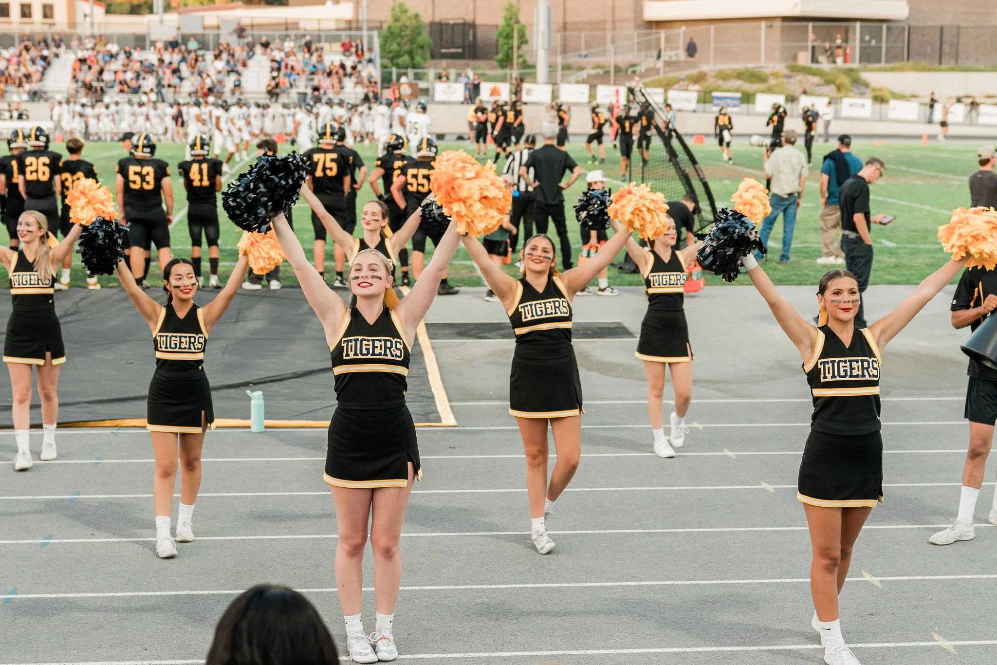 Cheerleaders in black and yellow uniforms cheer during a football game, with football players, cheerleaders, spectators, and a football field in the background.