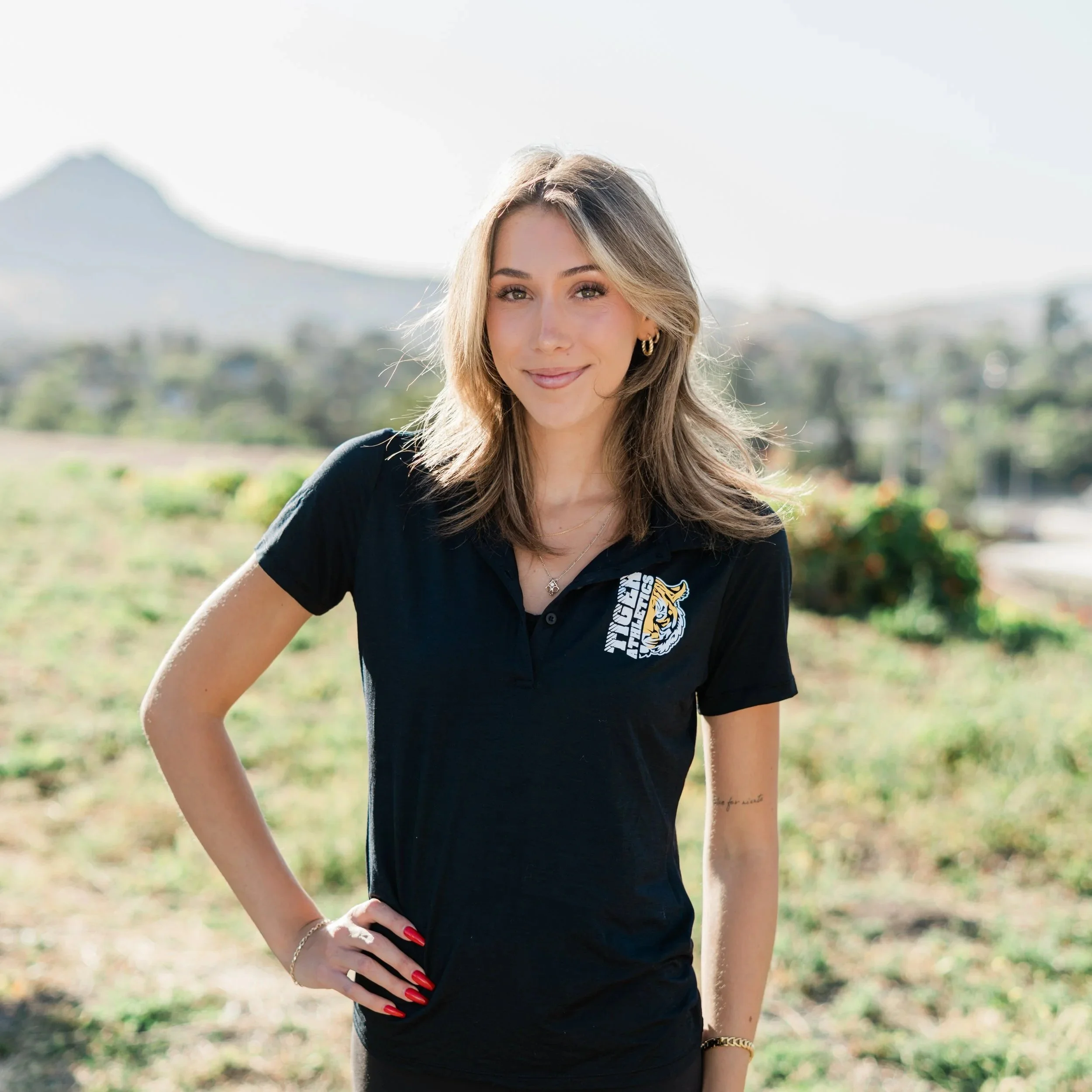 A young woman standing on a grassy field at a sports stadium, smiling at the camera with sunlight behind her. She is wearing a black polo shirt with a tiger logo and the word 'Tigers' on it, black leggings, and a delicate bracelet.