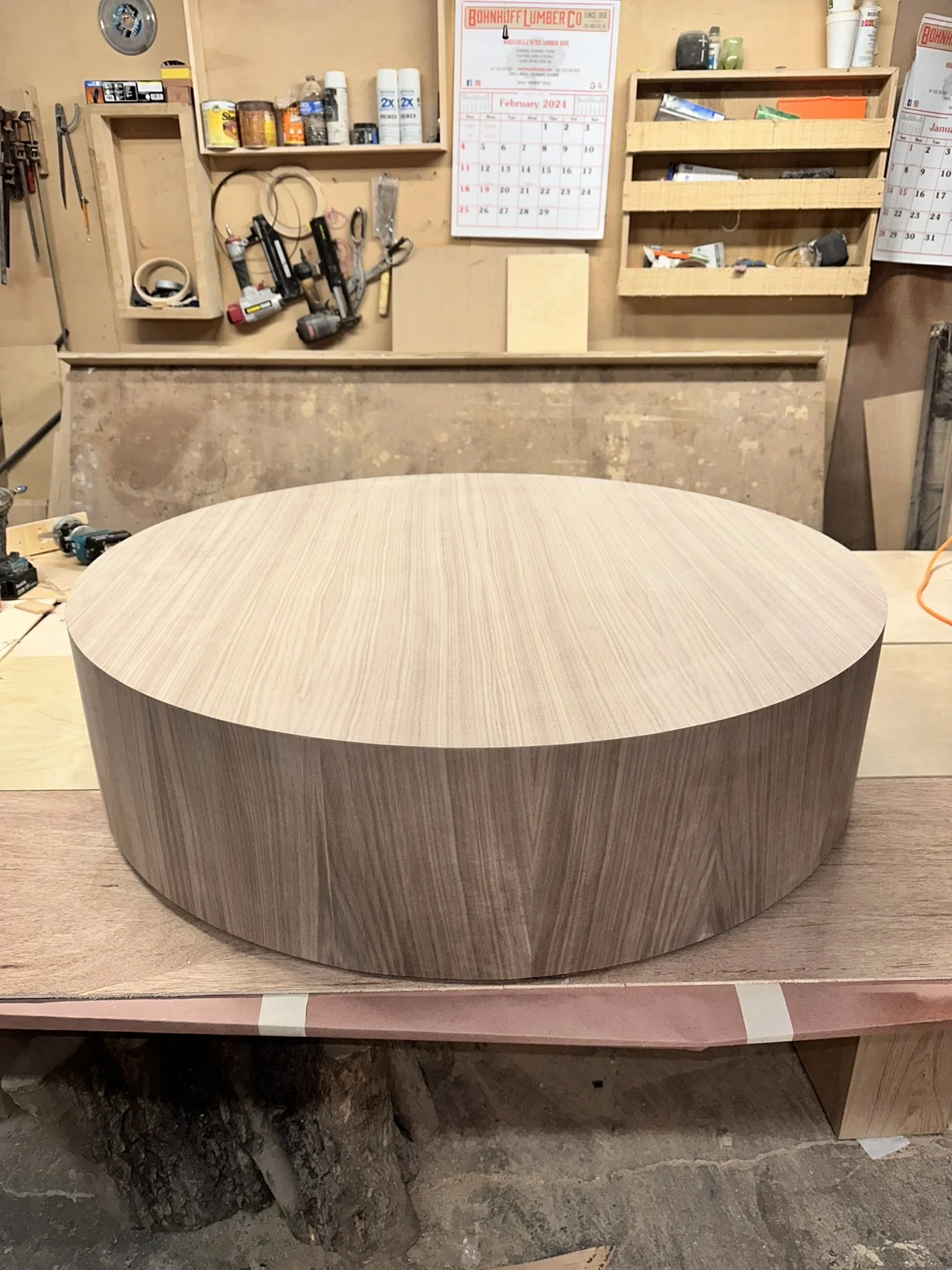 A rounded wooden table in a woodworking workshop, with tools and supplies on the workbench and shelves in the background.