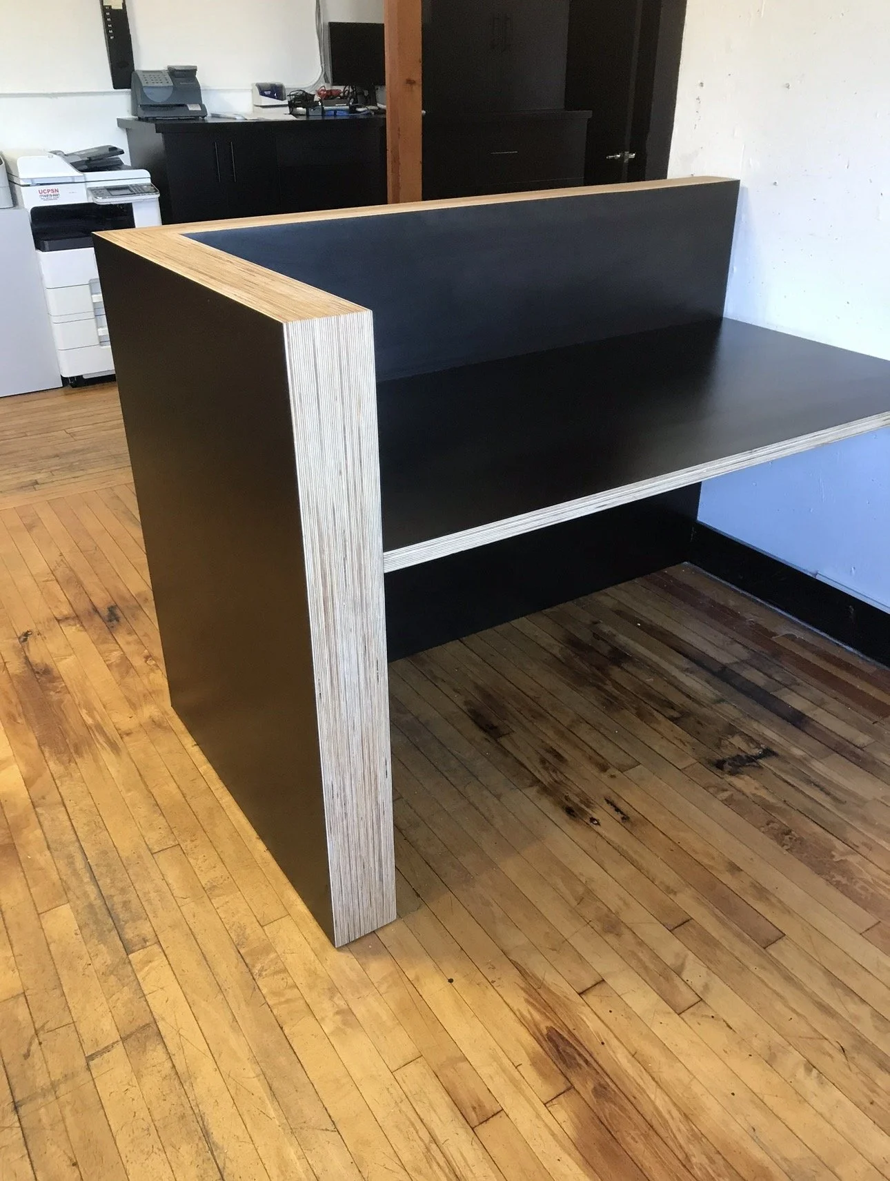 A modern wooden and black desk in an office setting with a wooden floor, in front of a white wall and office equipment in the background.