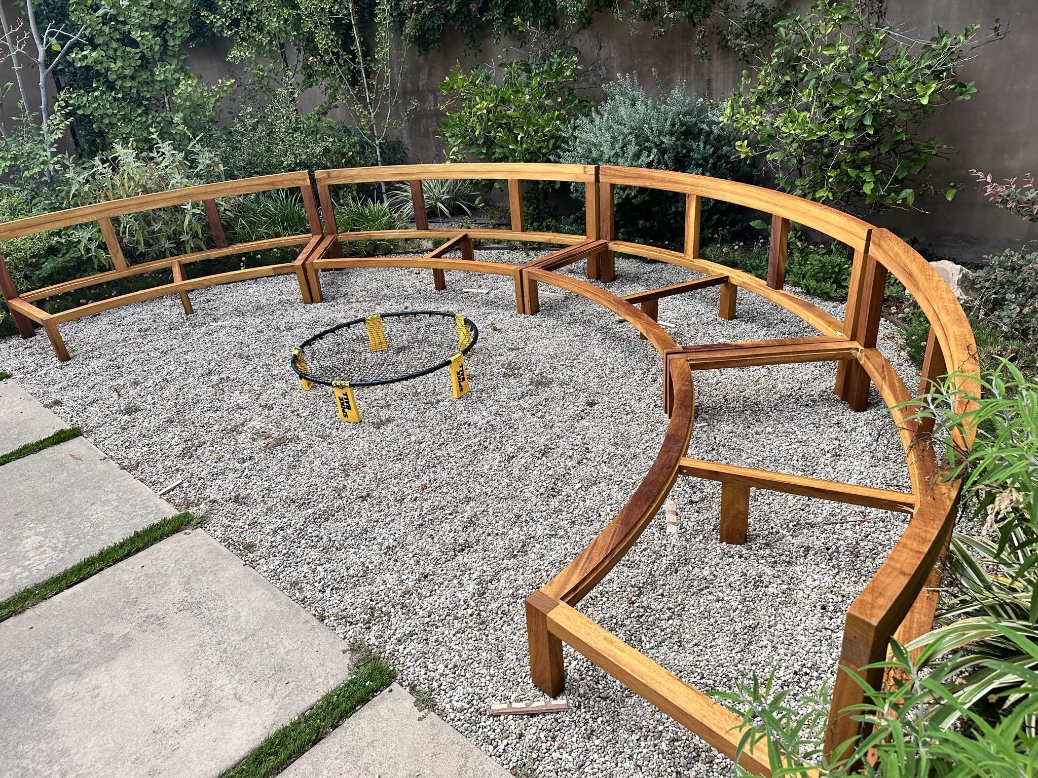 A wooden outdoor bench frame on gravel ground in a backyard garden with green bushes and trees in the background.