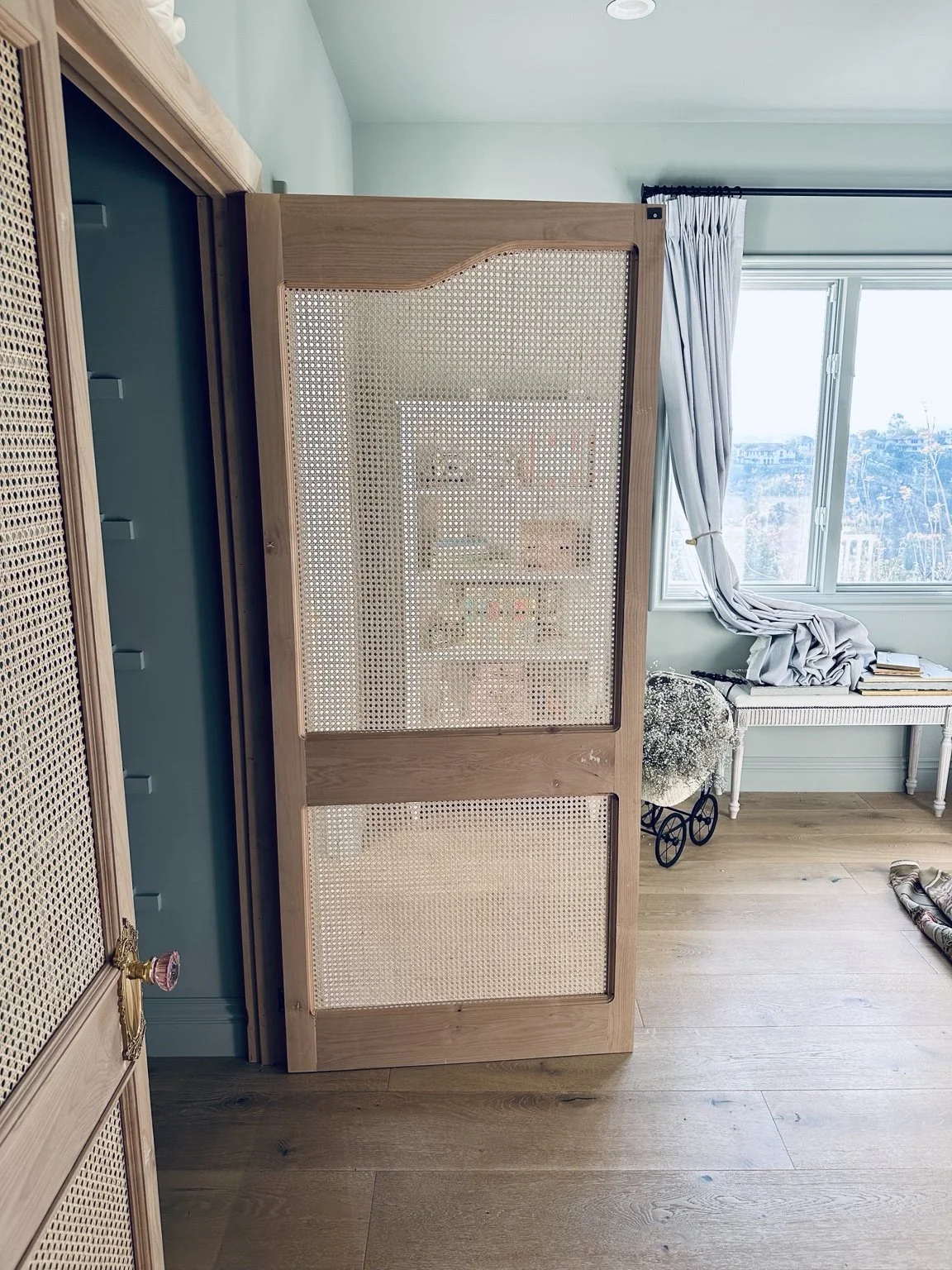 A wooden room divider with a woven pattern, positioned in a room with a hardwood floor, near a window with light blue curtains. A white table with books and a flower arrangement is nearby.