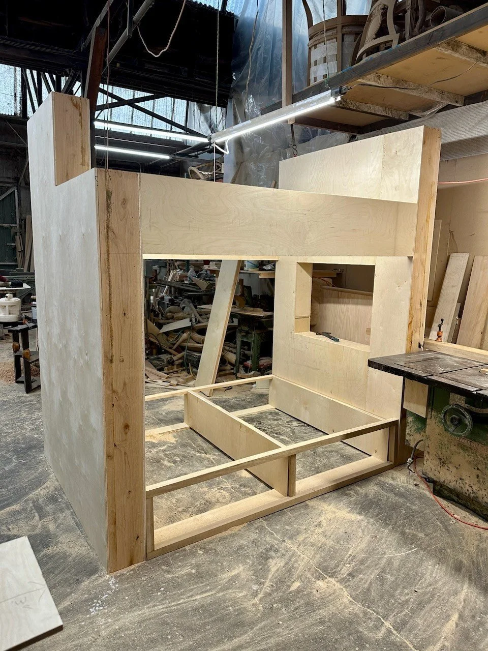 Wooden structure under construction in a workshop, with partly assembled frames and plywood panels.