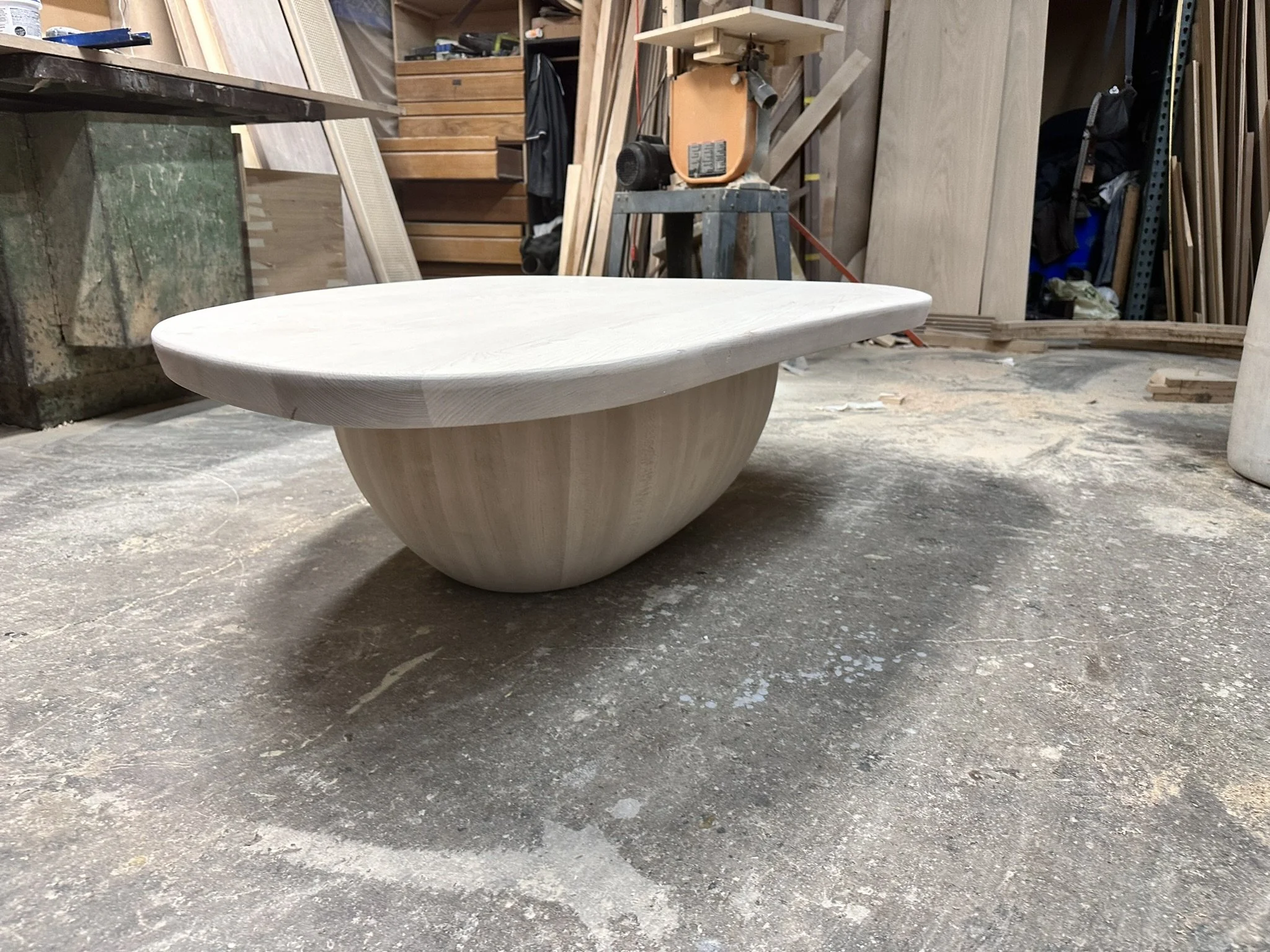A round, white wooden table in a woodworking workshop with tools and wood planks in the background.