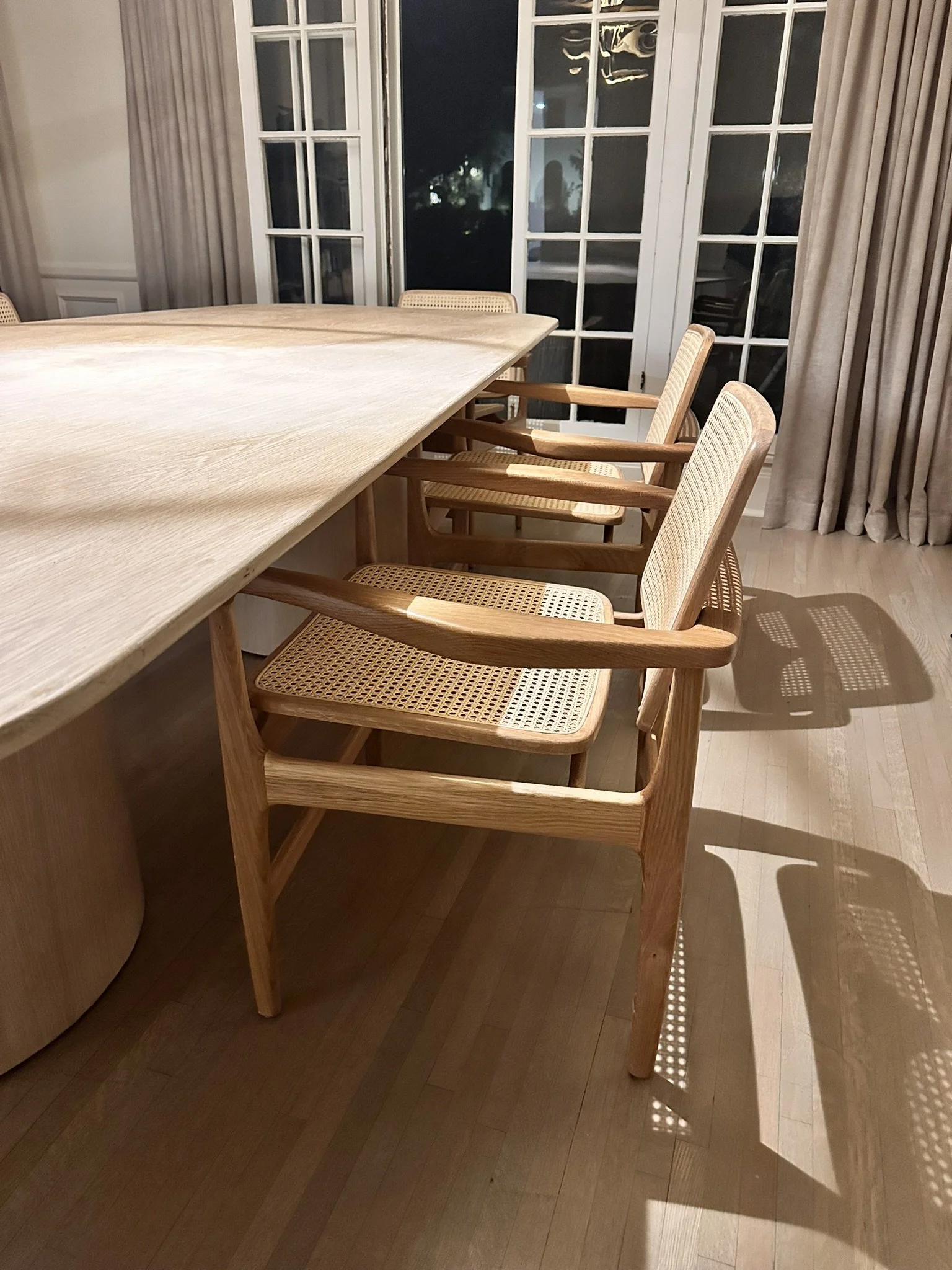 A wooden dining table with three wooden chairs featuring woven cane seats and backs, positioned near a window with glass panes and beige curtains, and a wooden floor underneath.