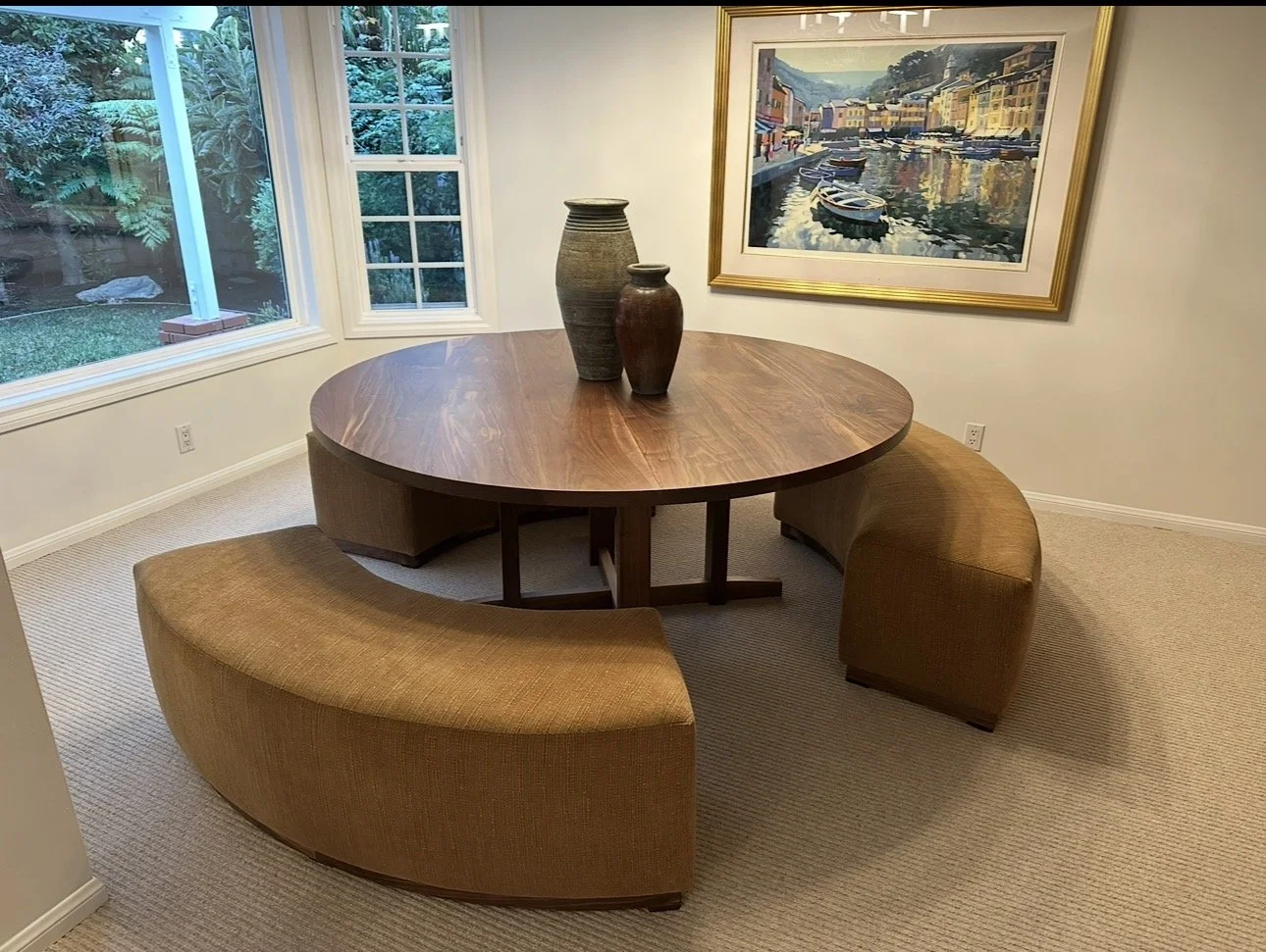 A round wooden dining table with three decorative vases in the center. The table is surrounded by curved upholstered benches. There is a large framed painting of a harbor scene on the wall, and windows showing greenery outside.