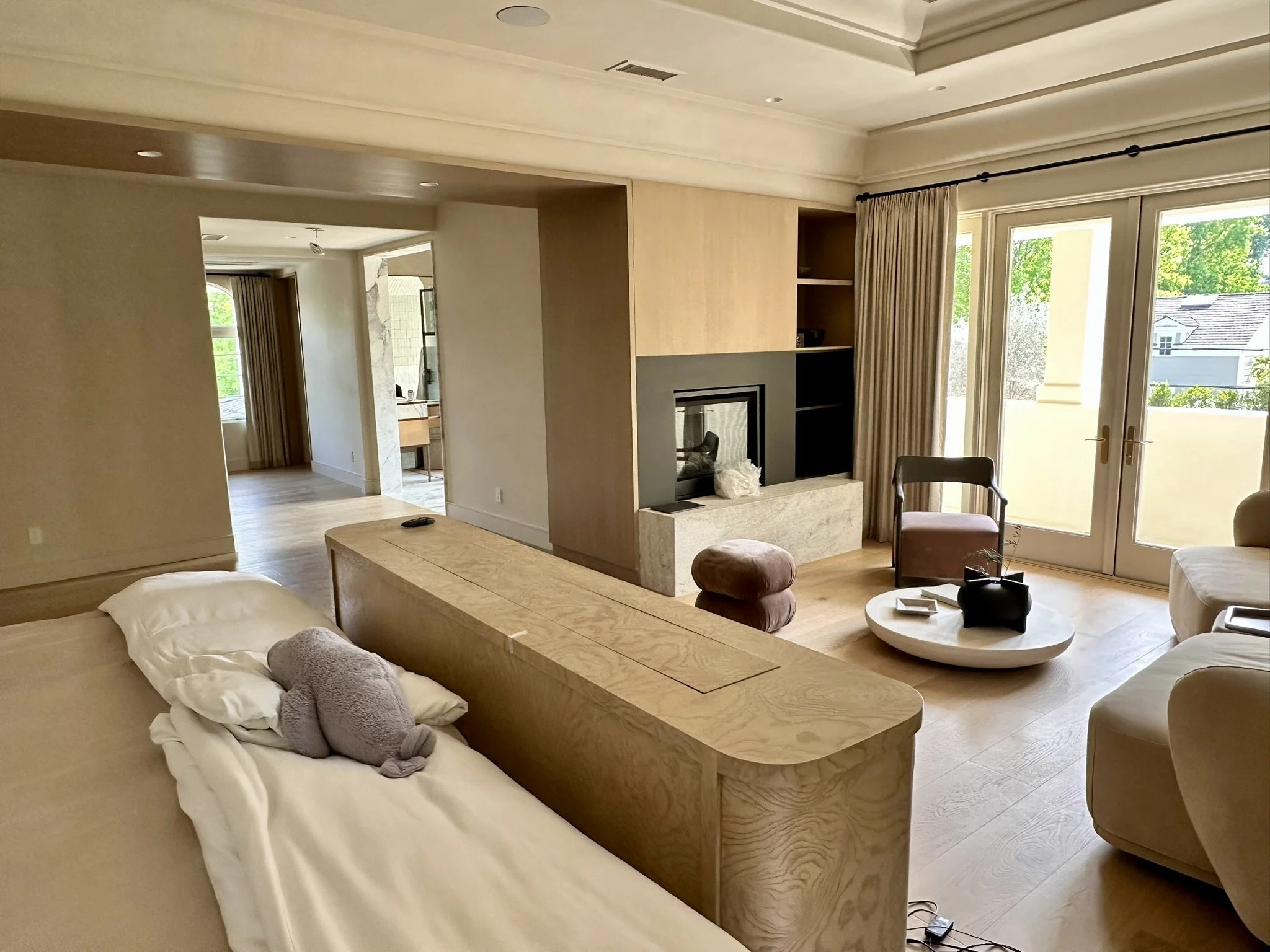 Living room with large windows, beige curtains, built-in fireplace, and wooden furniture, including a table with a black vase and plush toys on a bed.