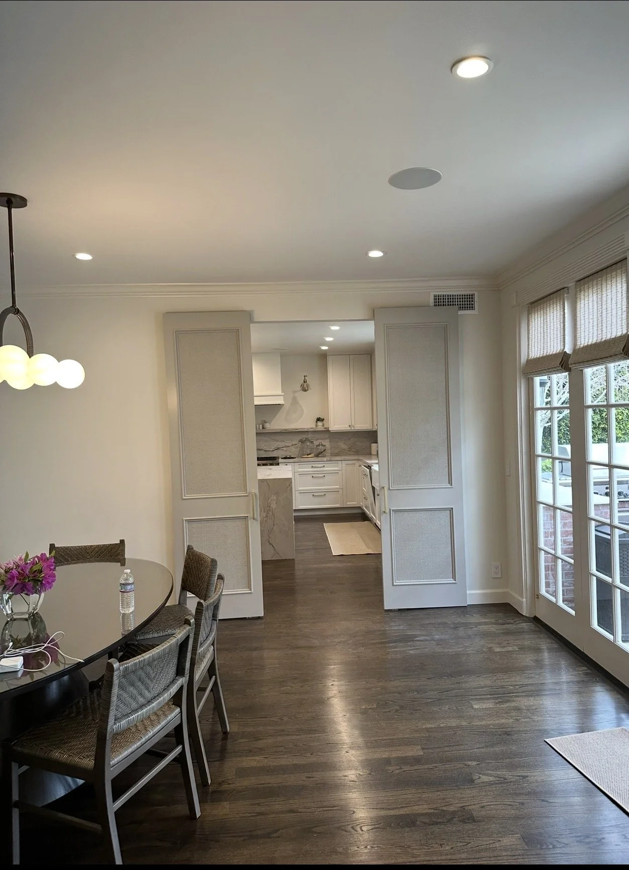 Open kitchen viewed from dining area with dark hardwood floors, white cabinets, marble backsplash, and large windows with beige shades.