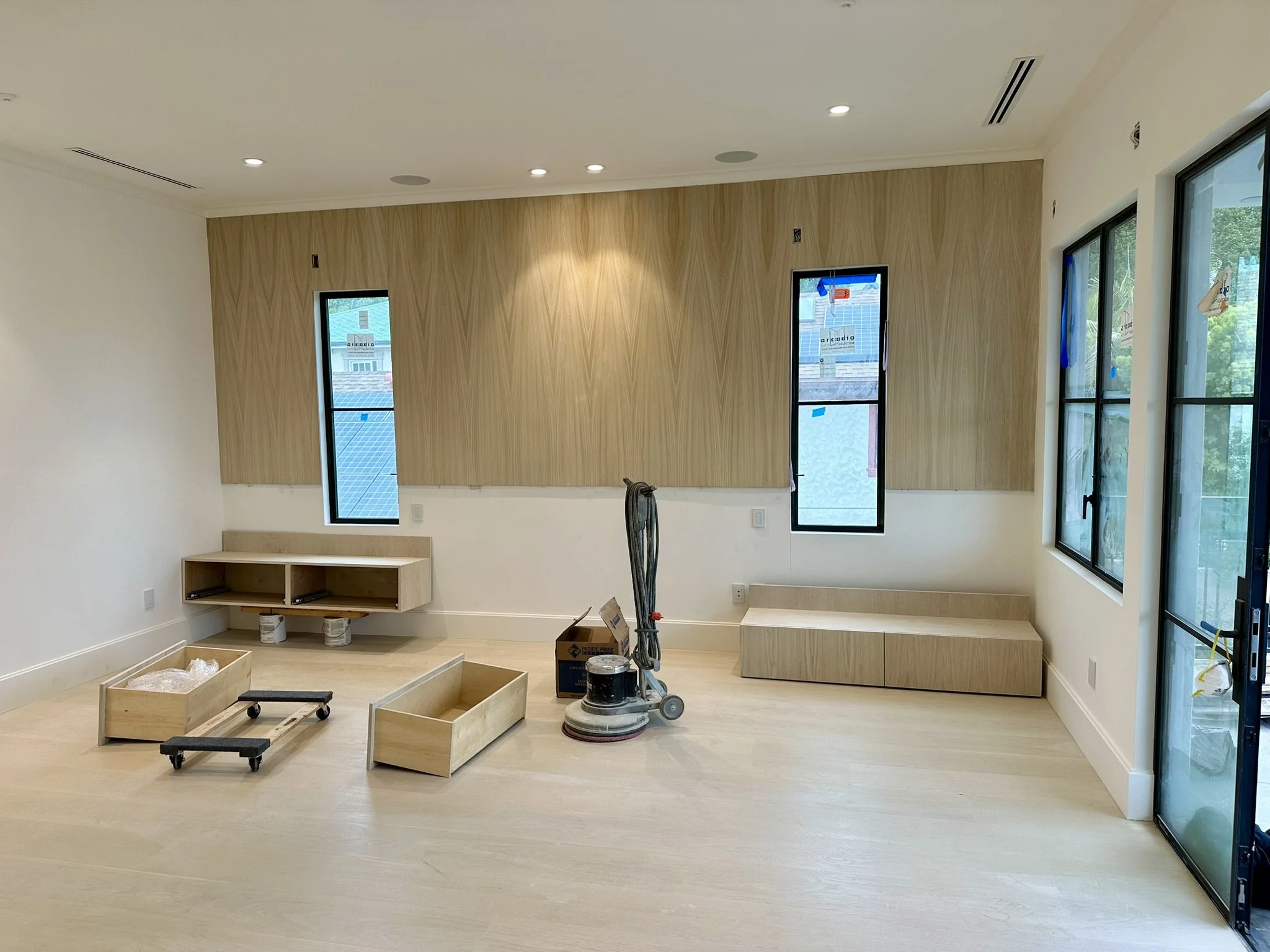 Interior room under construction with light wood flooring, beige walls, and a wood-paneled wall with three narrow windows. Construction tools and wooden boxes are on the floor, and a vacuum is also present. Large glass sliding door on the right with 