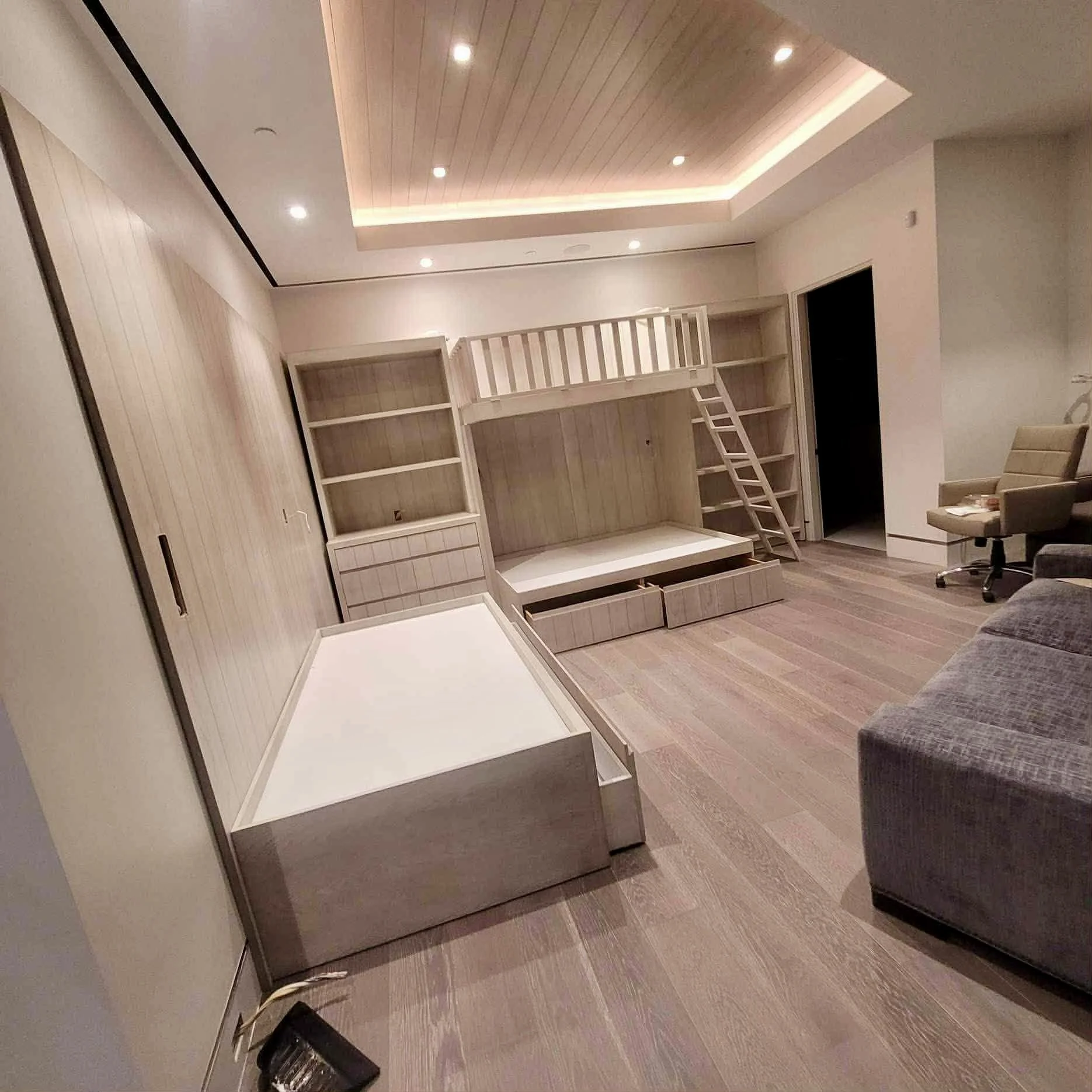 Modern living room with a built-in wooden desk, bookshelf, and loft bed with ladder. The room has recessed ceiling lights and hardwood flooring.