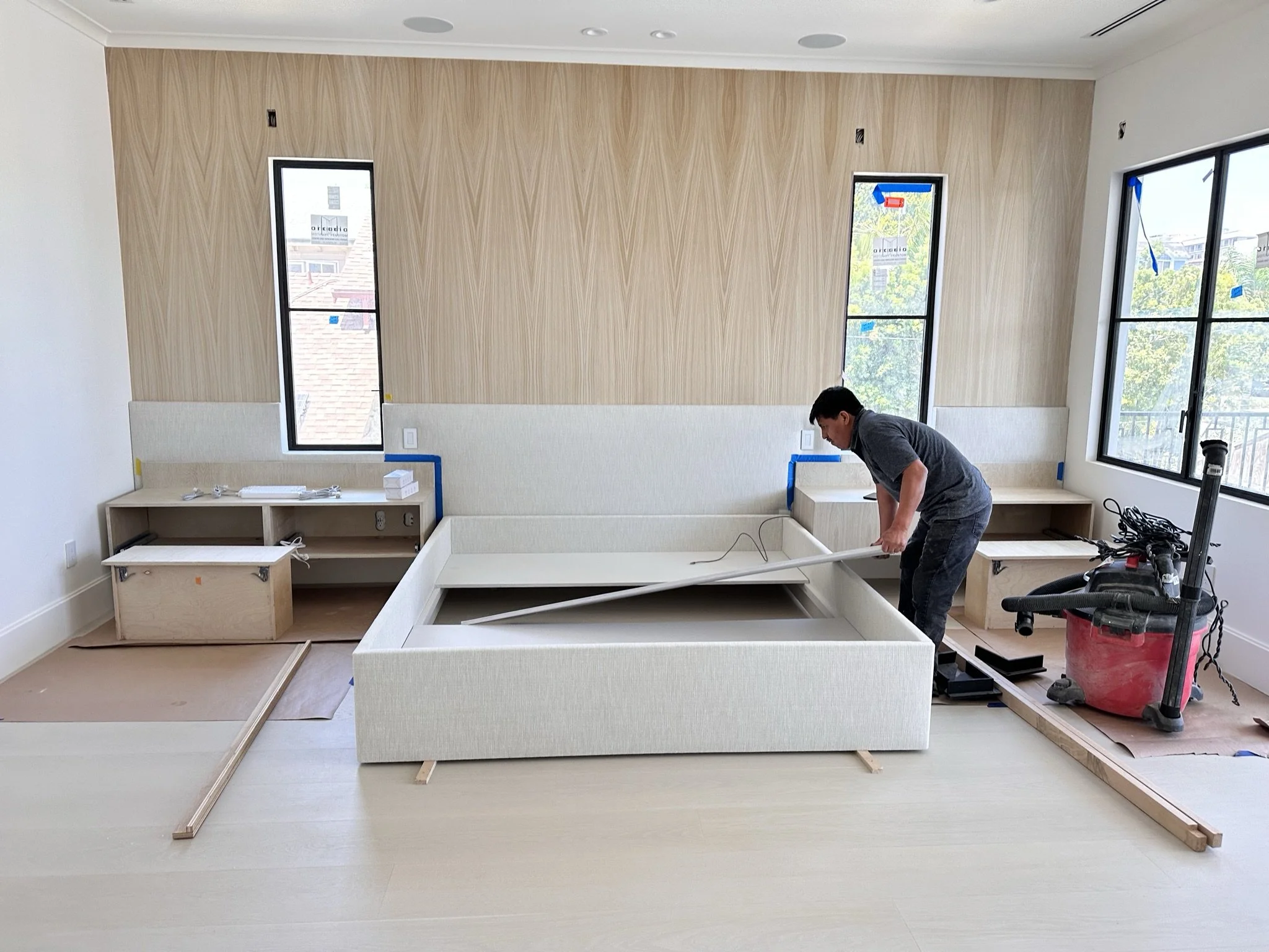 A person is installing or assembling furniture in a room with large windows and light-colored walls. The furniture appears to be a bed or seating with a built-in frame, surrounded by tools and construction materials.