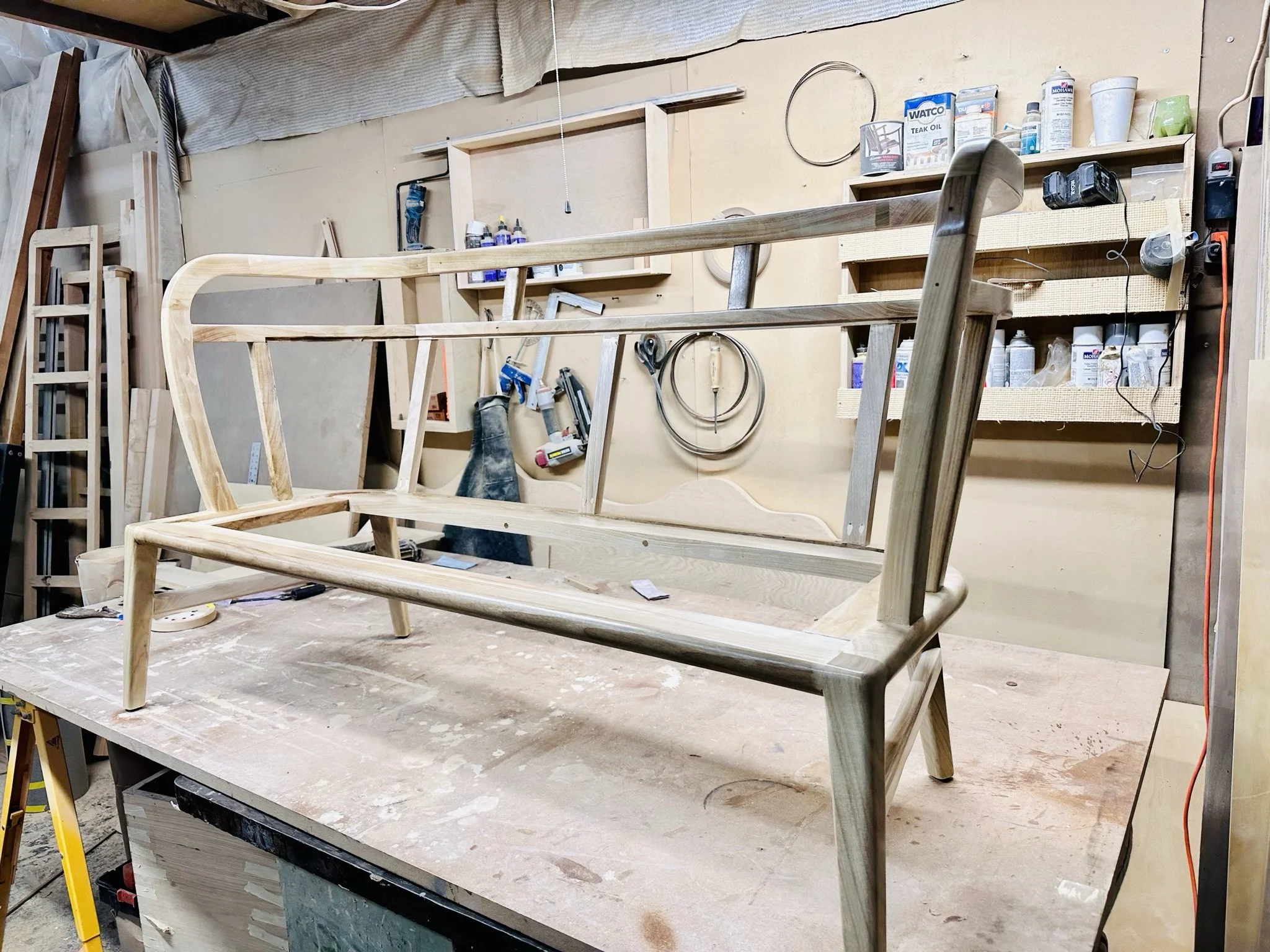 Wooden chair frame in a woodworking shop with tools and supplies visible in the background.
