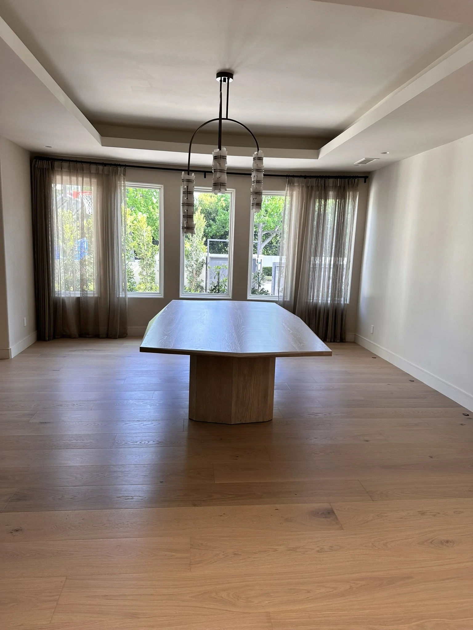 A dining room with a wooden table, large windows with sheer curtains showing greenery outside, and a modern chandelier hanging from a tray ceiling.