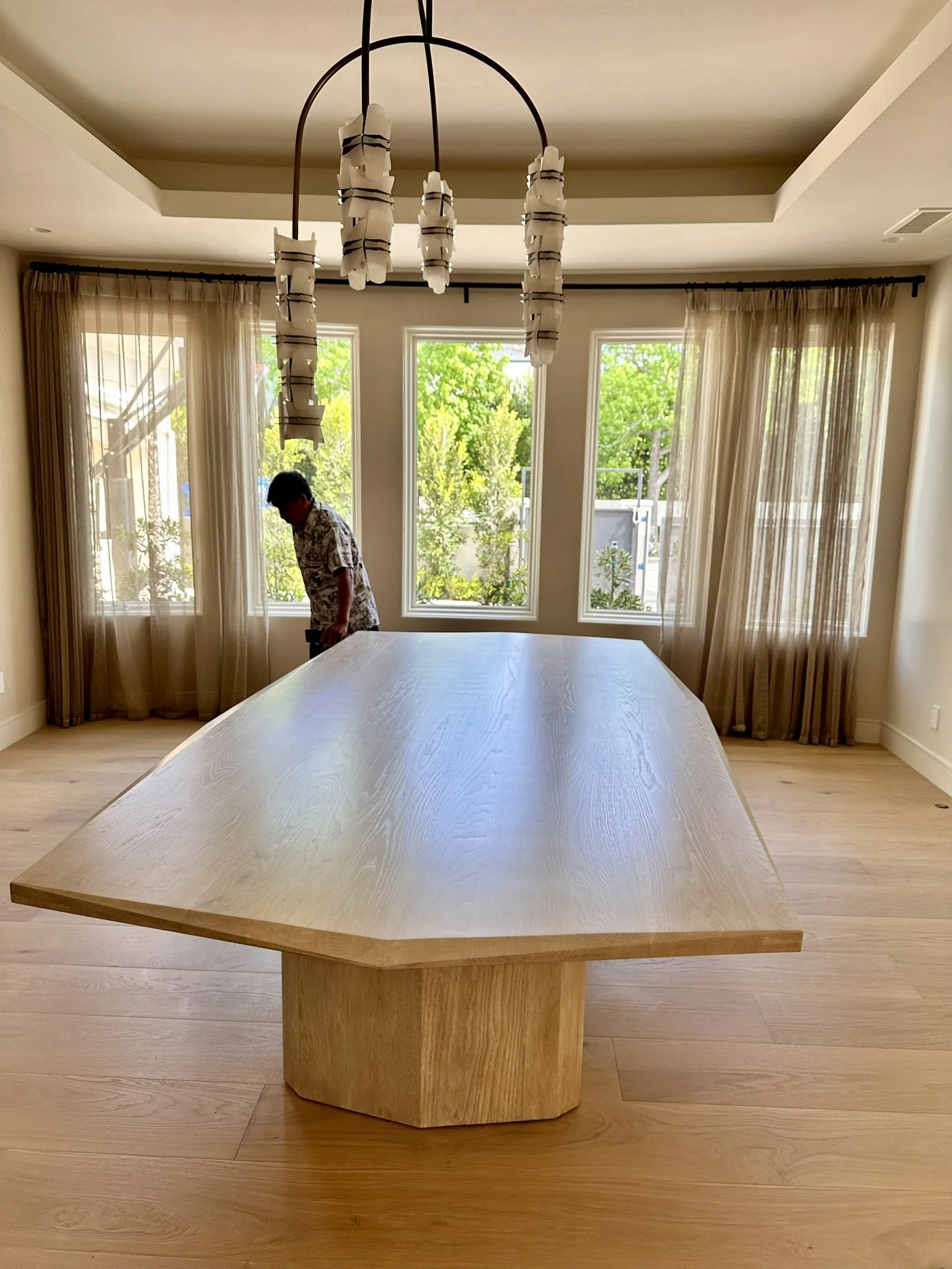 Interior view of a room with a large wooden dining table, beige curtains on three windows with green trees outside, and a modern chandelier hanging from the ceiling. A person is standing near the window in the background.