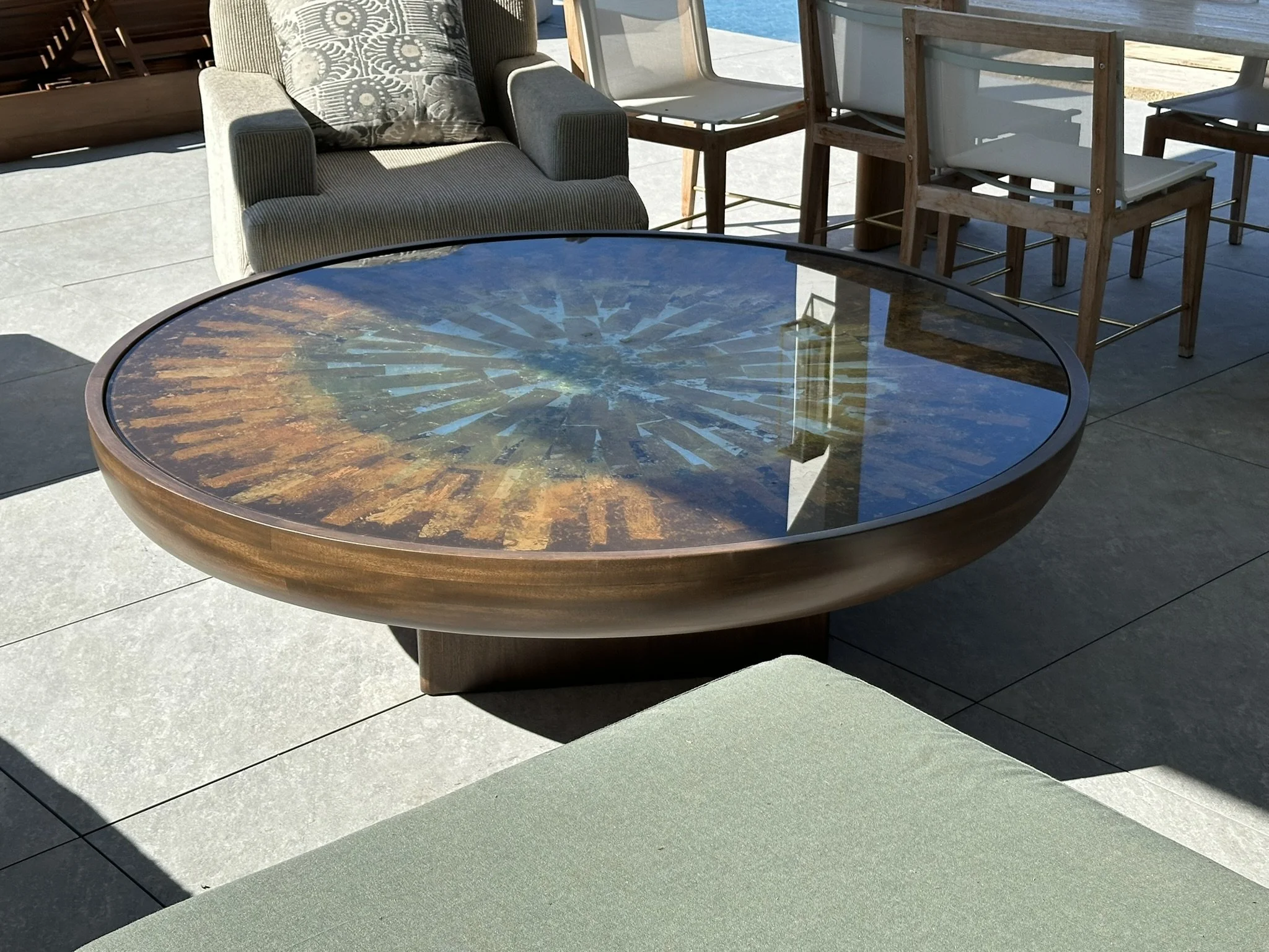 Outdoor patio area with a reflective glass-top table, an upholstered armchair with a patterned pillow, several wooden chairs with white cushions, and a swimming pool in the background.