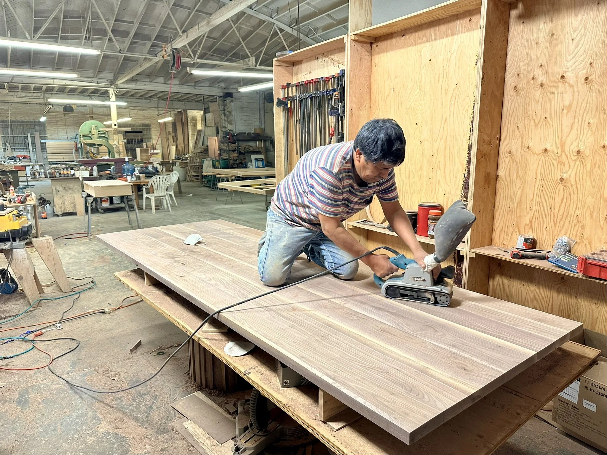 A man working on a woodworking project in a workshop, cutting a large wooden panel with a power saw. The workshop is filled with tools, woodworking equipment, and unfinished projects.