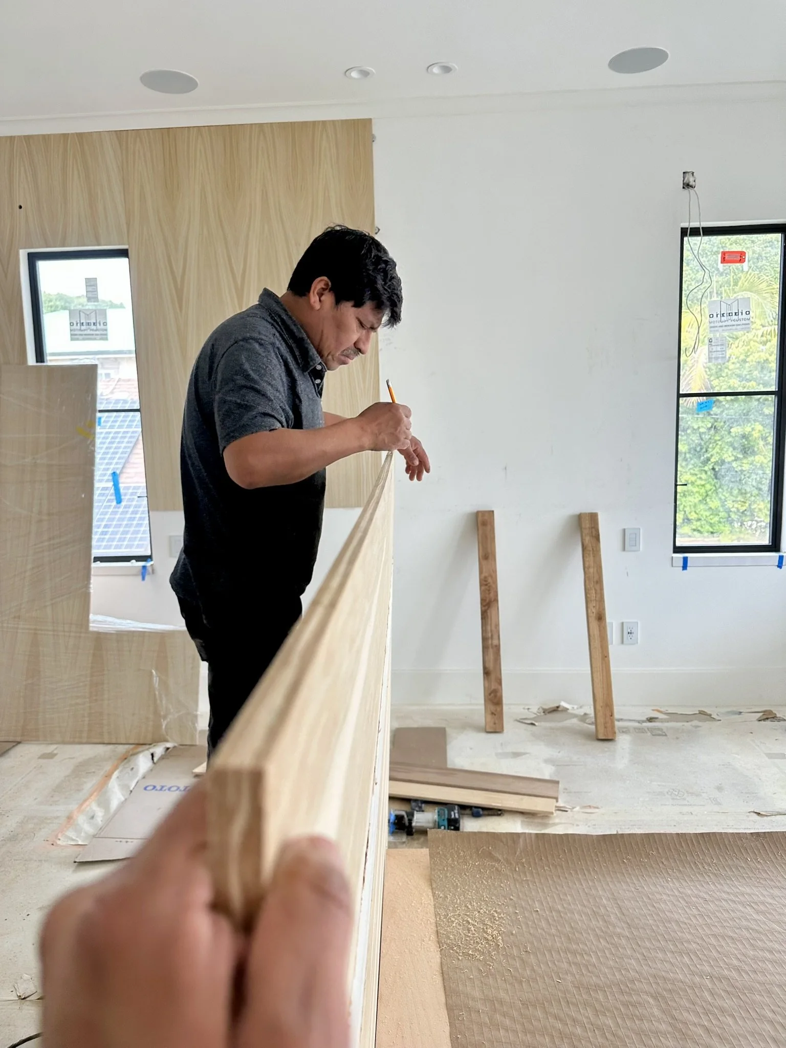 A man in a dark shirt is measuring a piece of wood indoors with a pencil in his hand. The room appears to be under construction, with wooden beams, tools, and materials on the floor. There are windows letting in natural light.