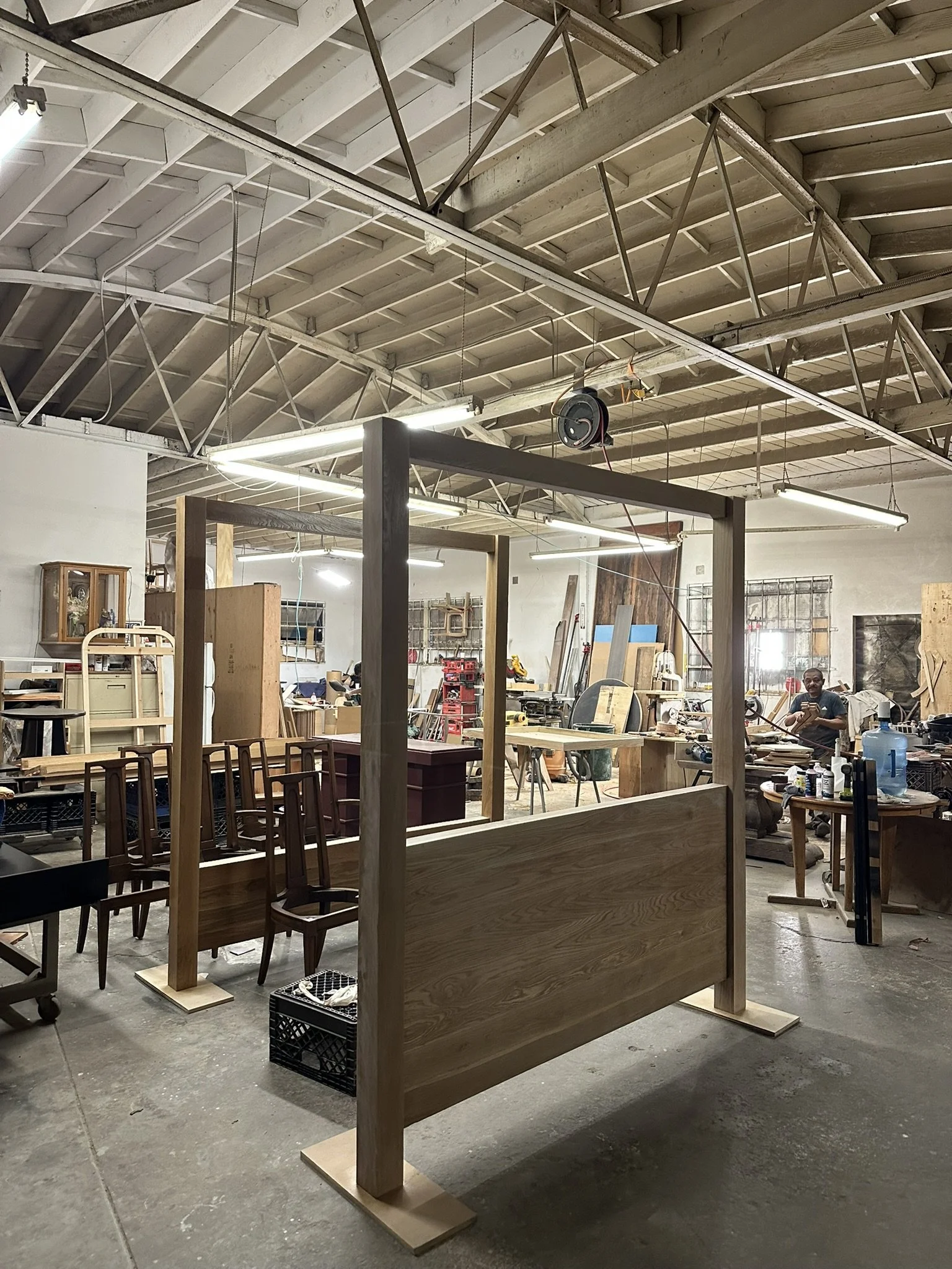 Workshop with wooden furniture pieces in progress, wooden frames, tools, and materials scattered around, a man working in the background, exposed roof with lighting.
