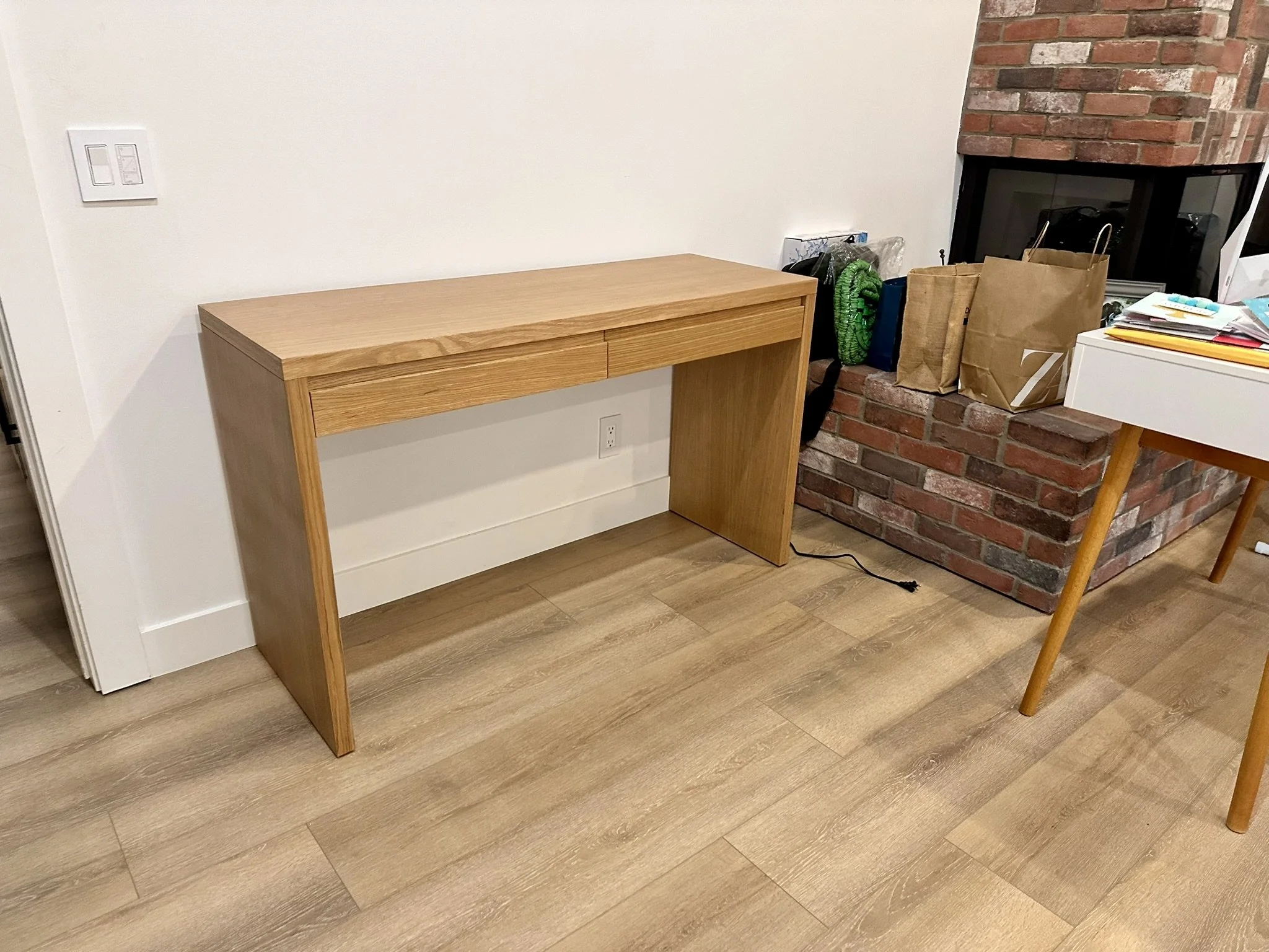 A wooden desk against a white wall with a light switch. To the right, there is a brick fireplace with a pile of papers, bags, and a green object on the hearth, and a partial view of a white table and a wooden chair.