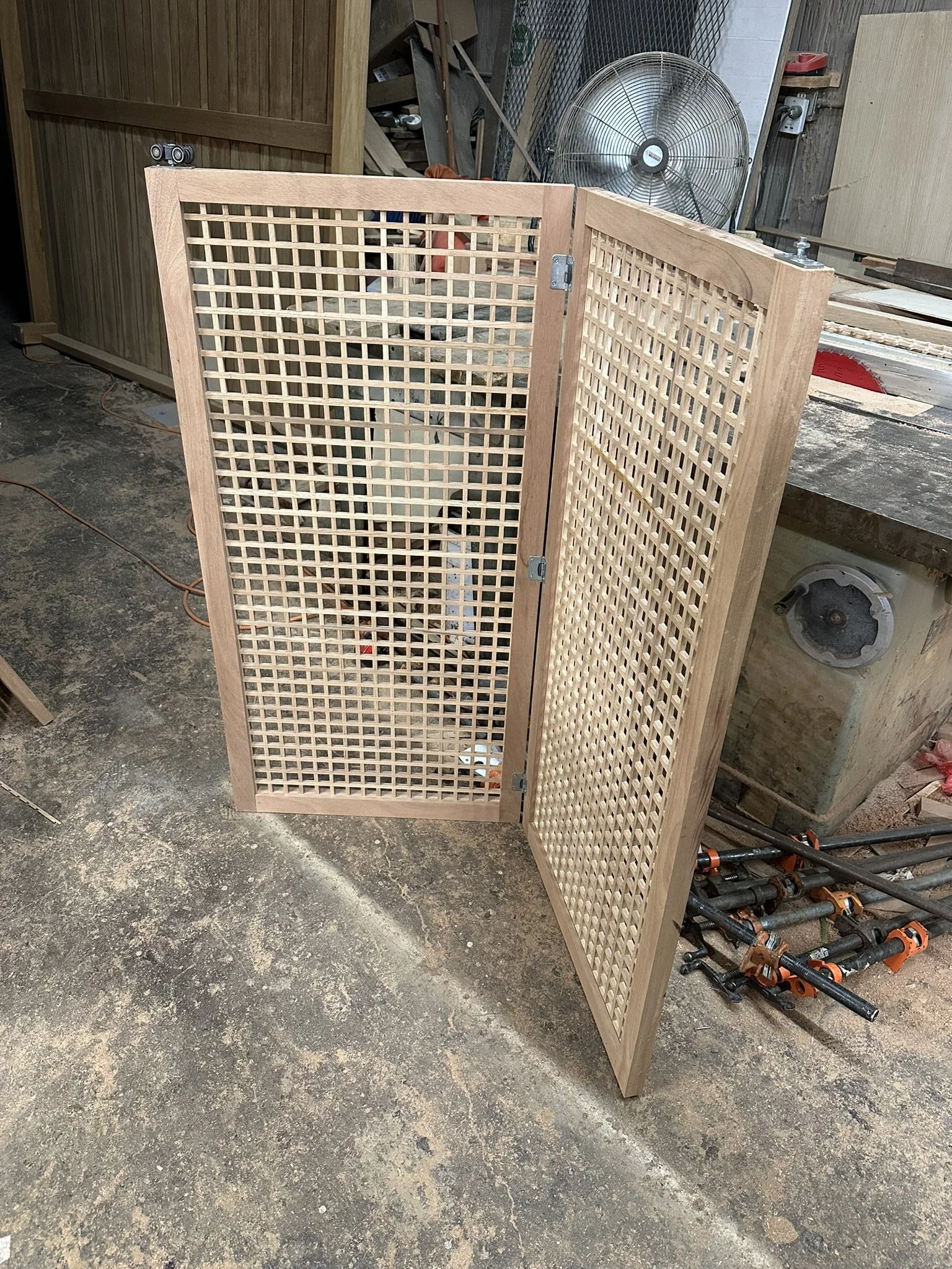 A partially assembled three-panel wooden folding screen with a wicker pattern in a woodworking workshop. The workshop has a fan, various tools, and wood pieces in the background.