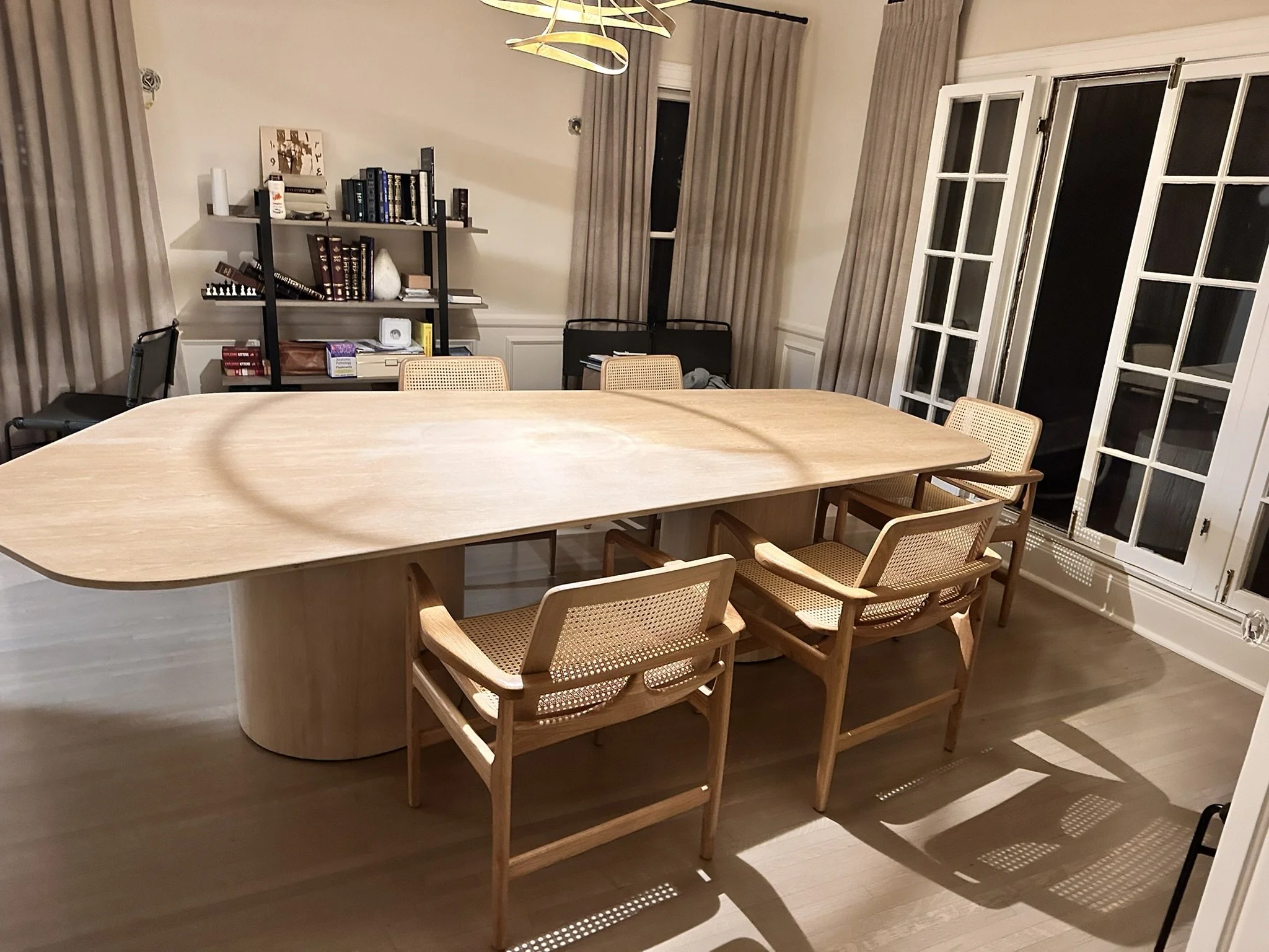 A dining room with a large oval wooden table surrounded by tan and black chairs. There is a black bookshelf with books and decorative items against the wall, and large windows with beige curtains. The floor is wooden, and there is a gold chandelier h