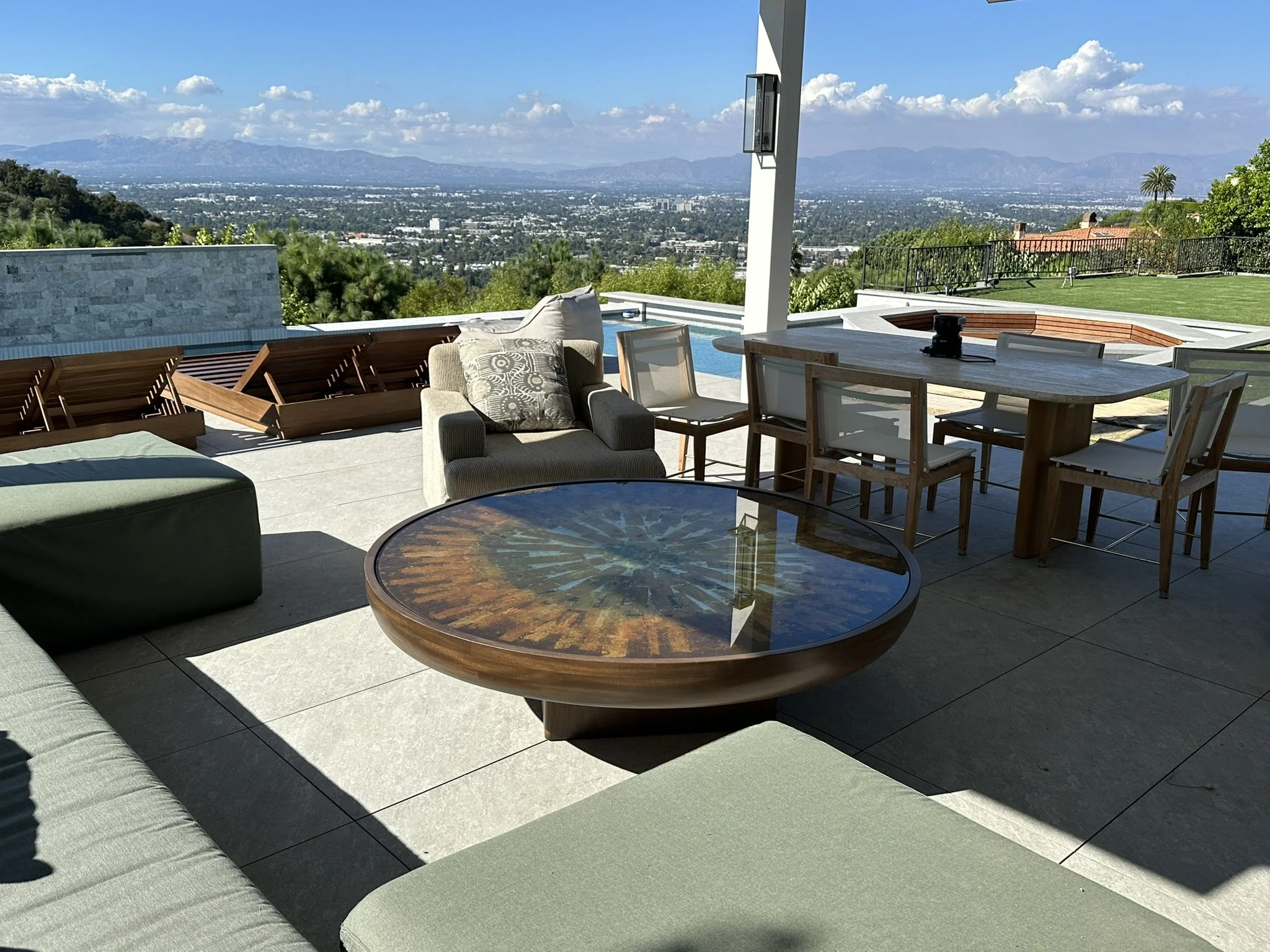 Outdoor patio area with a view of a city and mountains in the distance. There are lounge chairs, a cushioned chair, a round table, and a dining table with chairs. The patio has a swimming pool and a grassy yard enclosed by a black fence.
