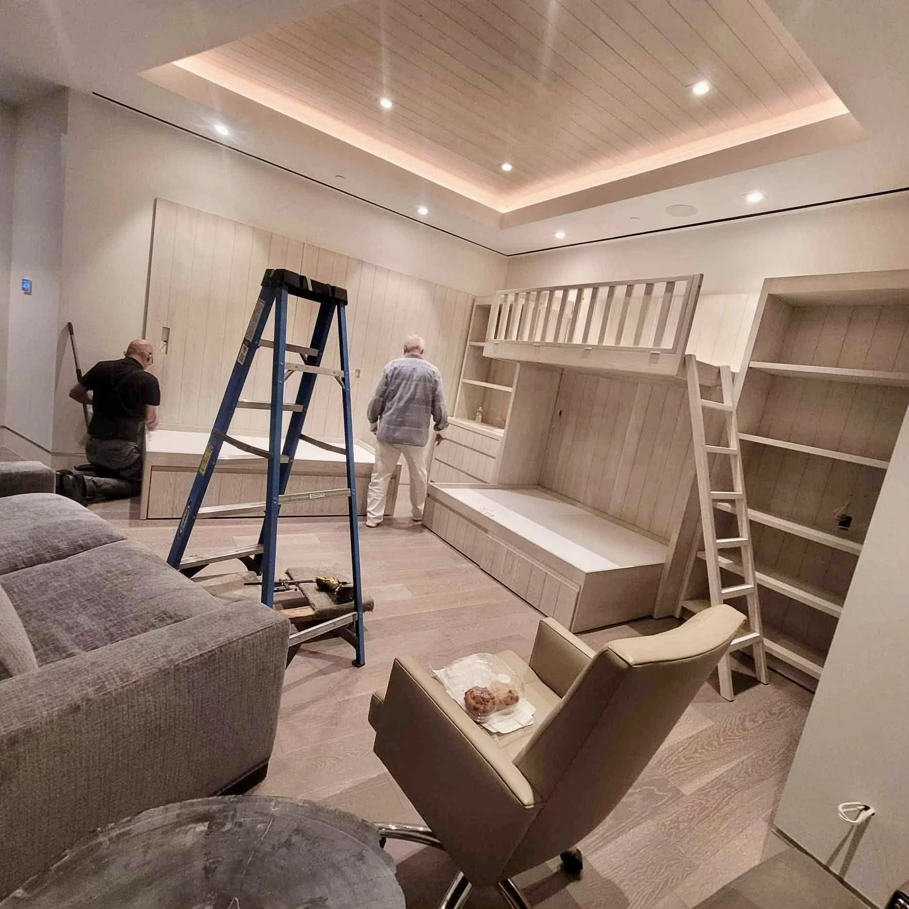 Interior of a room with a high ceiling, lights, and unfinished wooden furniture including a bunk bed and shelves, with two men working on the furniture.