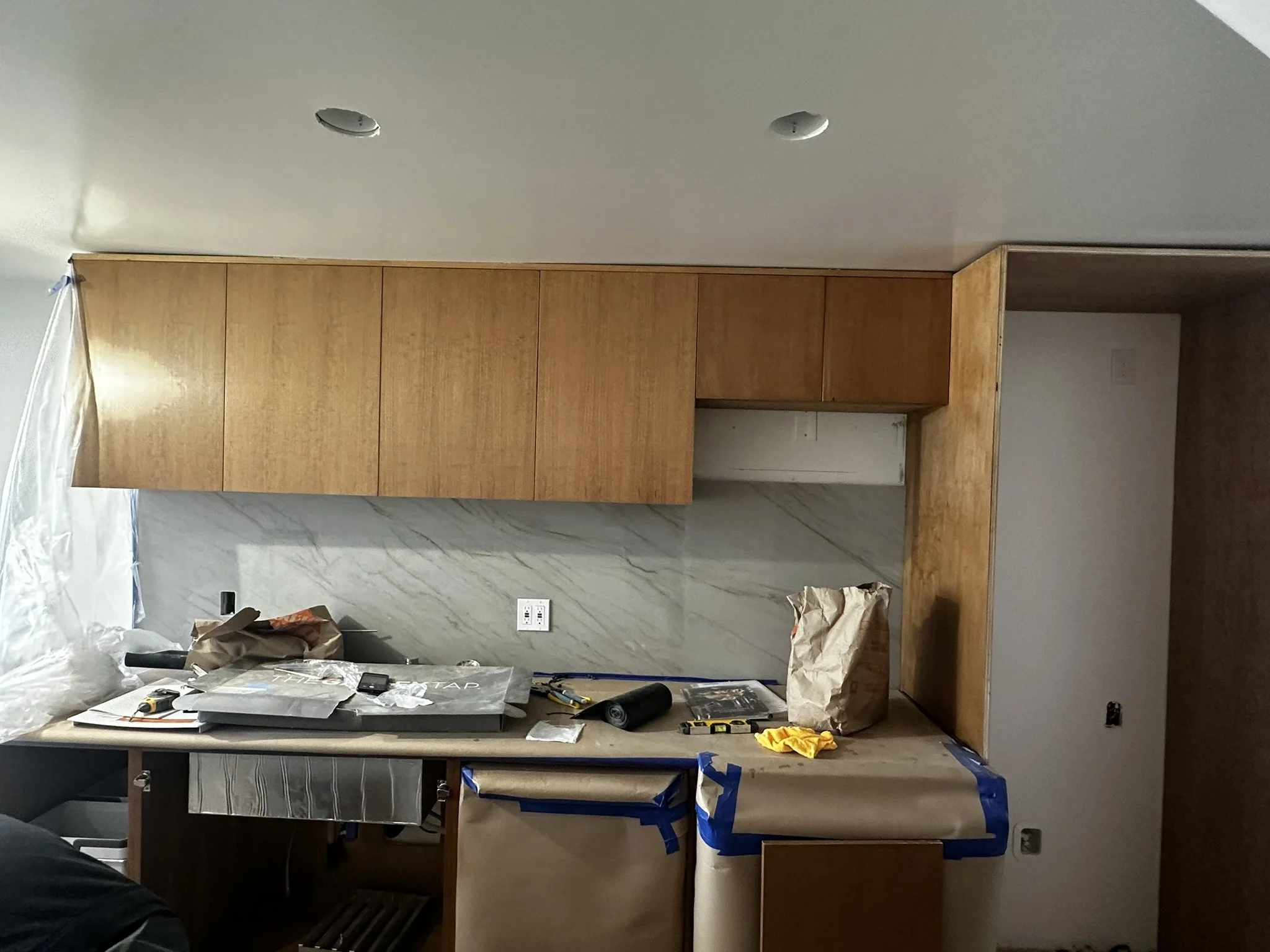 Interior of a room under construction with wooden cabinets, tools, and supplies scattered on a table, and plastic coverings protecting surfaces.
