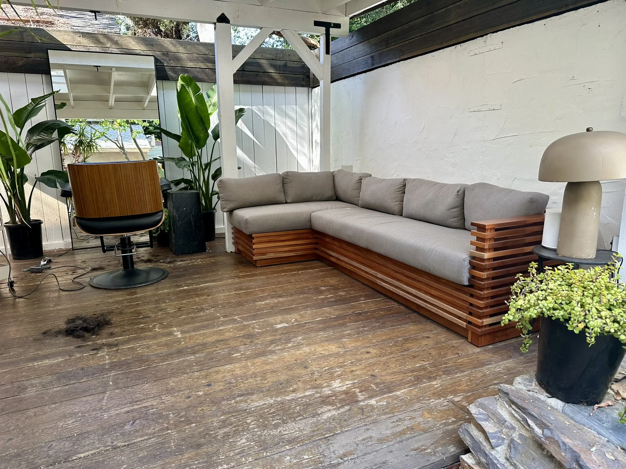 A living room corner with a beige sectional sofa, a modern table lamp, tall green houseplants, a round mirror, and wooden flooring. There is a white wall and a dark wooden roof structure. A mirror reflects part of the outdoor area with trees.