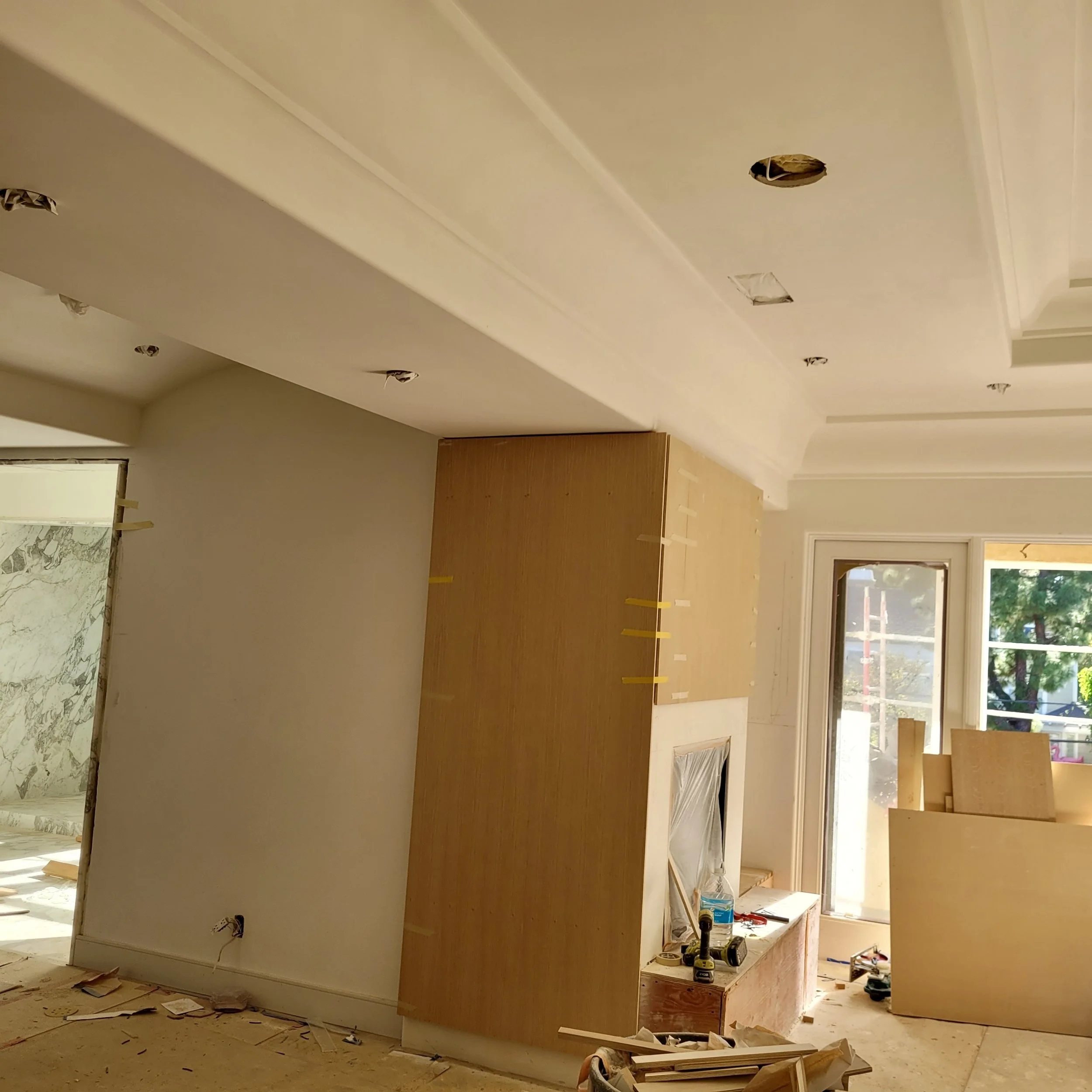 Interior of a house under renovation, with drywall installed and light fixtures being prepared. Construction tools and materials are scattered on the floor, and the ceiling has holes for lighting fixtures.