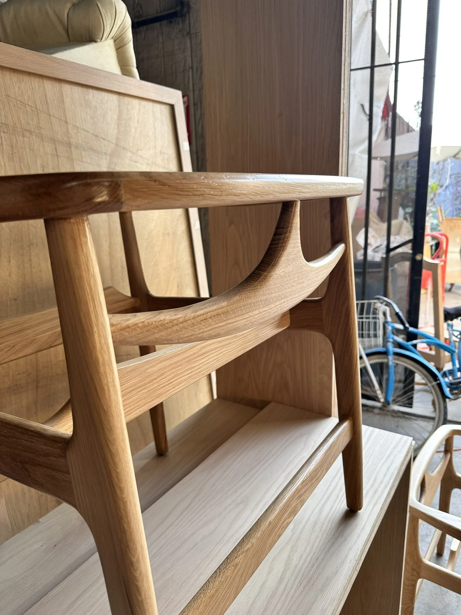 Rows of wooden furniture, including chairs and shelves, positioned in a store or shop, with a window showing outside view of bicycles and outdoor scenery.