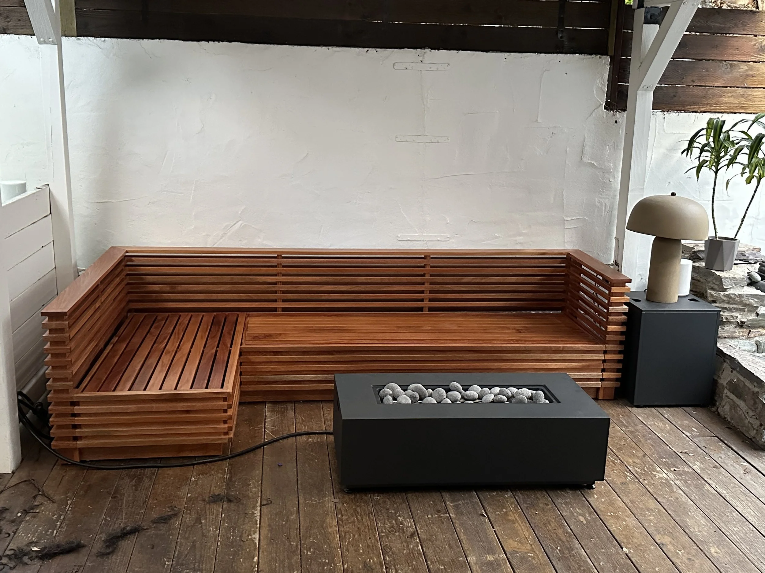 Outdoor seating area with a wooden bench, a small table with rocks on it, a white wall, and a potted plant.
