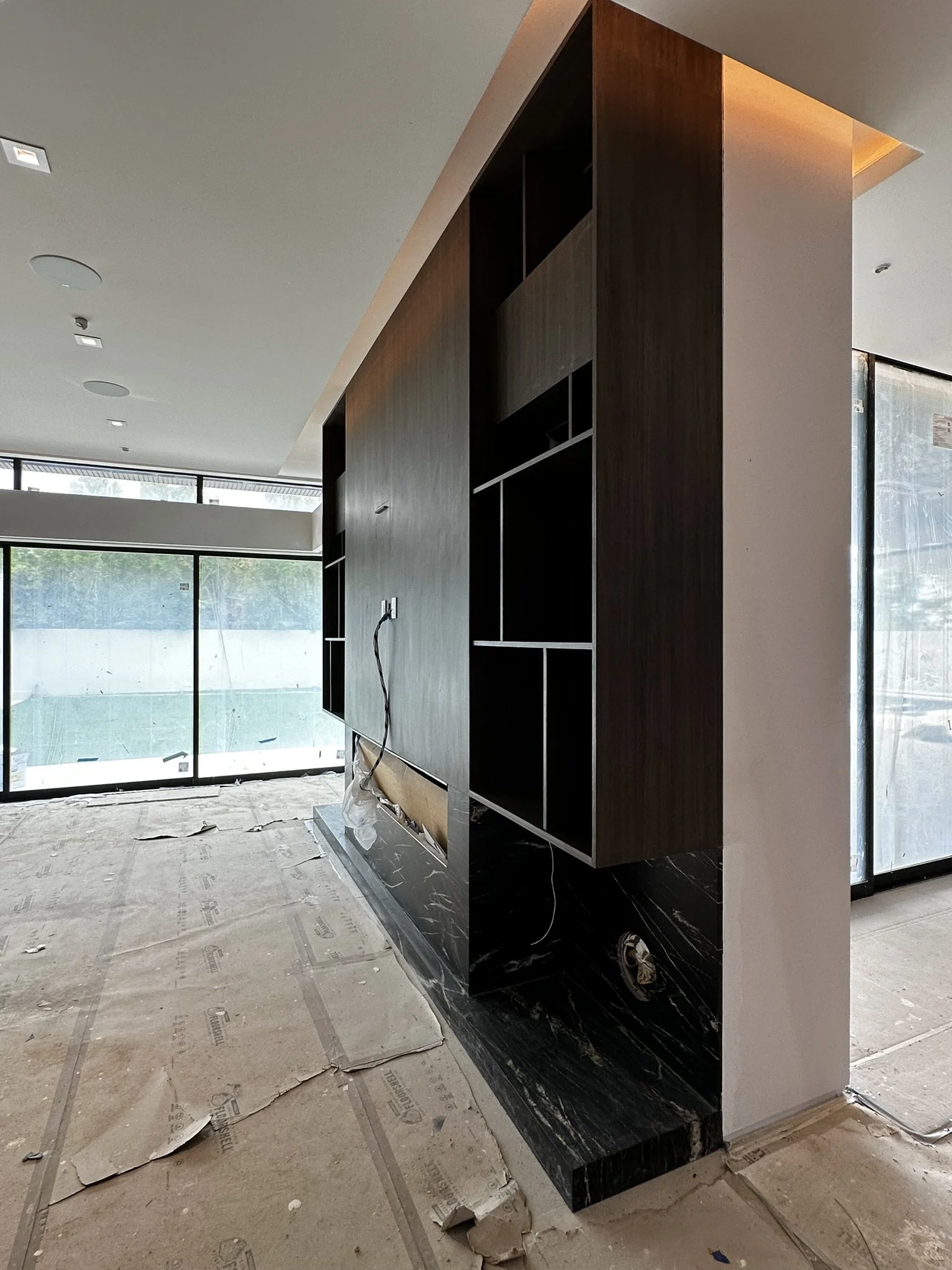 Interior of a room under construction, featuring a black and wooden built-in shelving unit with open cubbies, large windows letting in natural light, and construction debris on the floor.