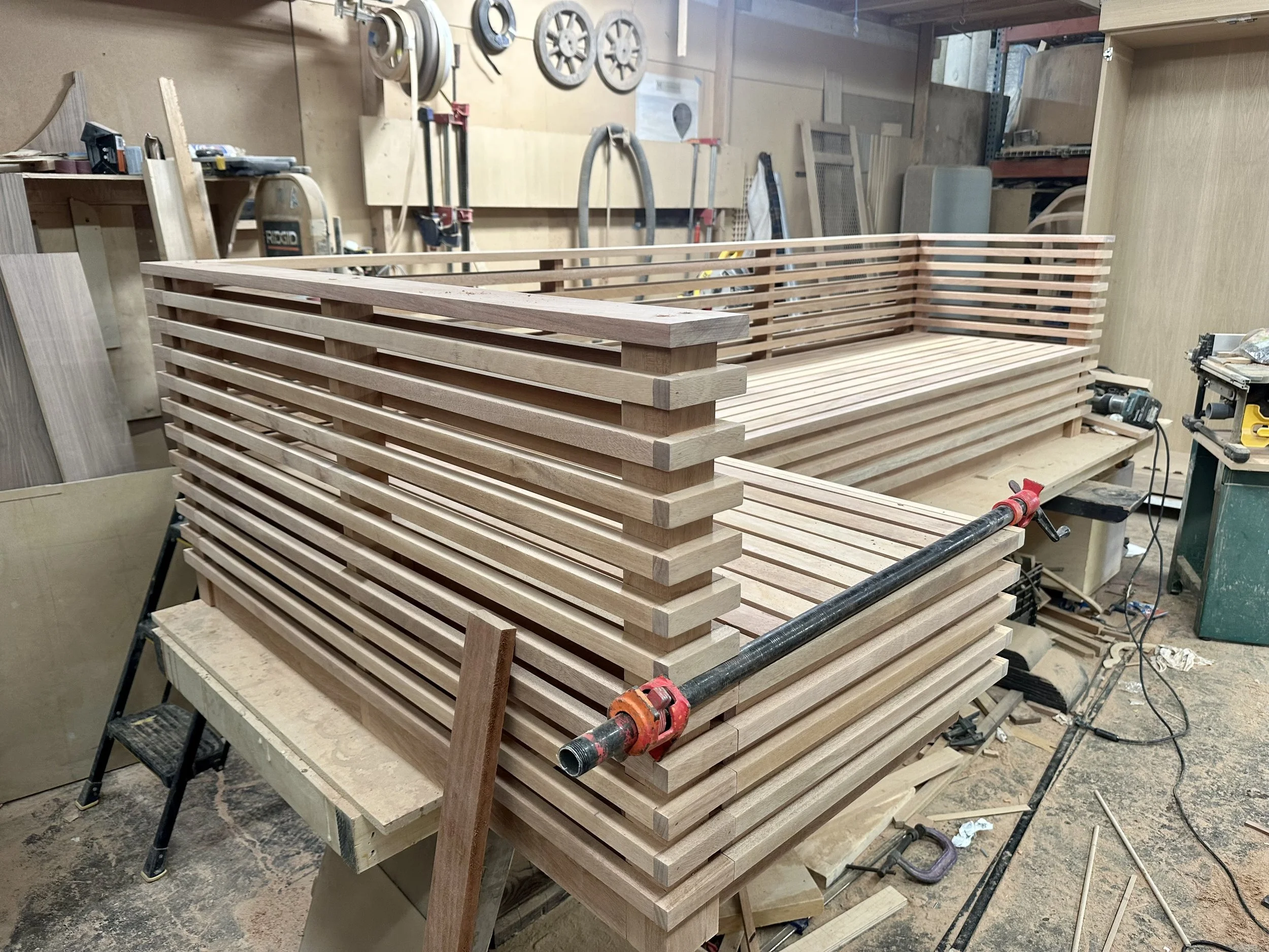 A woodworking workshop with a partially assembled wooden slat bench on a workbench, surrounded by woodworking tools and equipment.